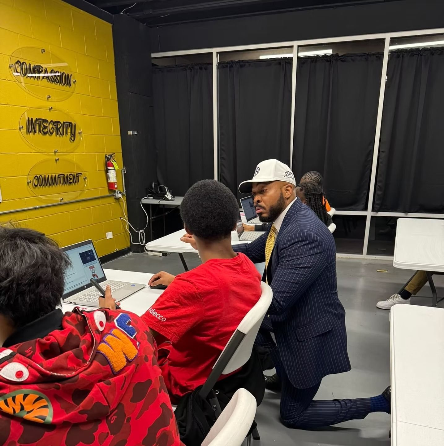 Man in suit assists student at a laptop in a classroom. Yellow wall with words 