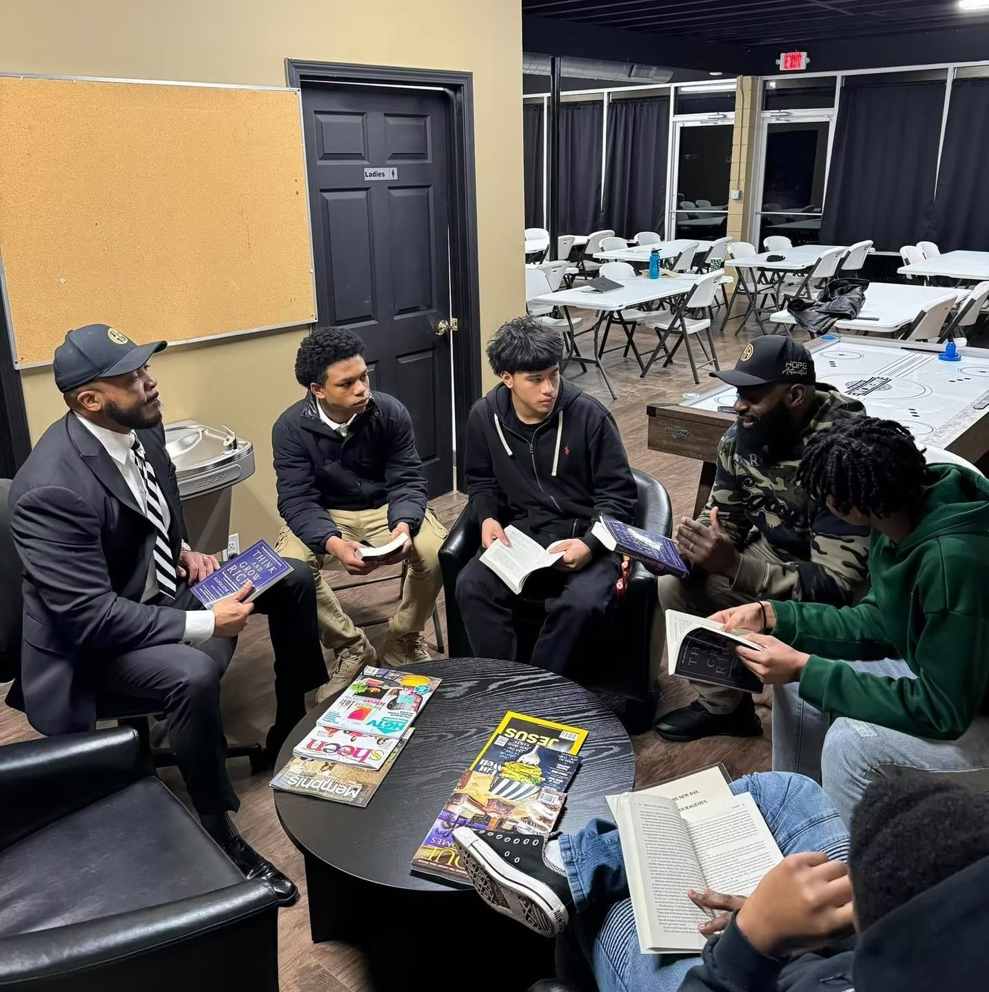 Group of people in a room, reading books. One person in a suit, others in casual clothing. Tables and chairs in the background.