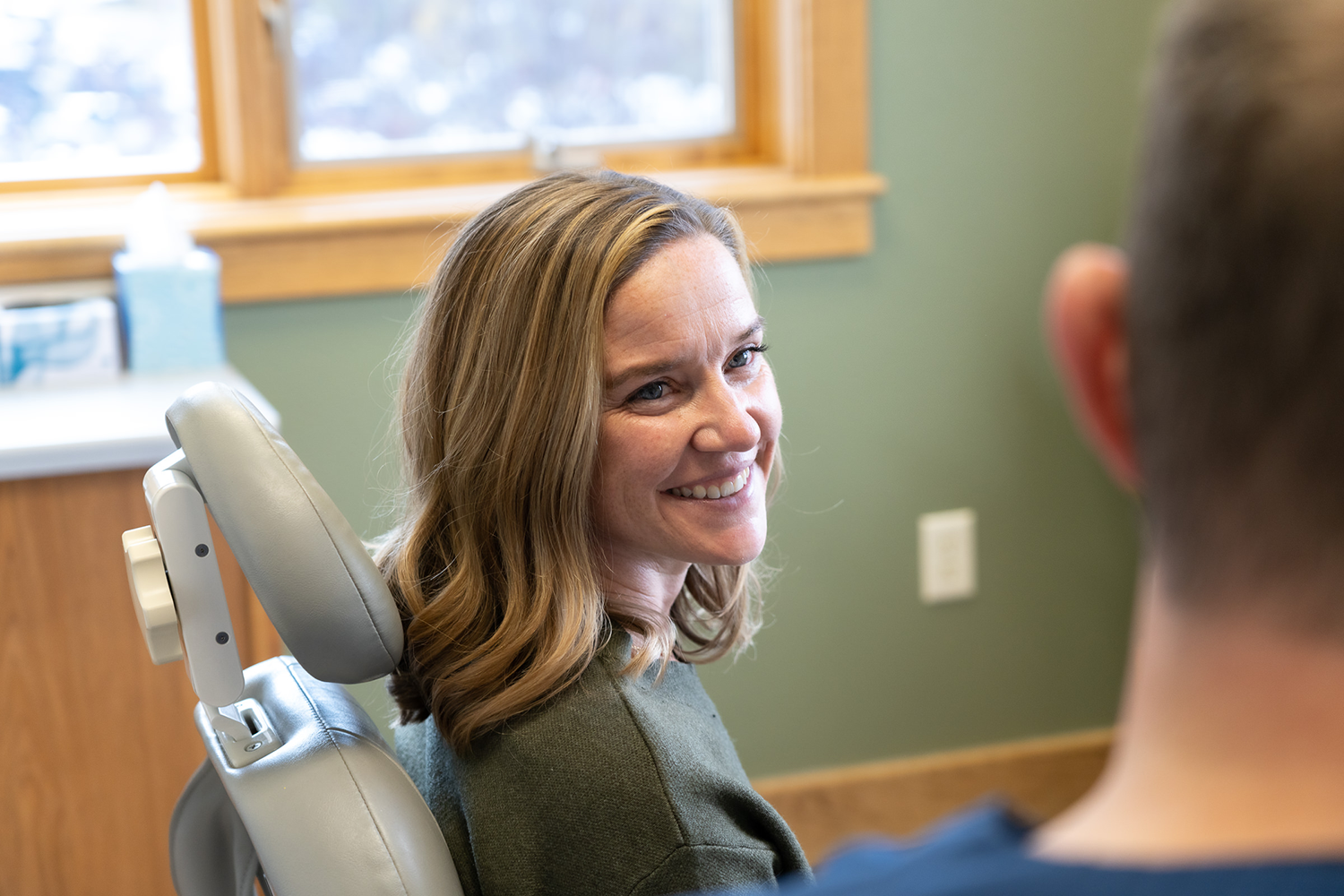 A woman is smiling while sitting in a dental chair talking to a dentist.