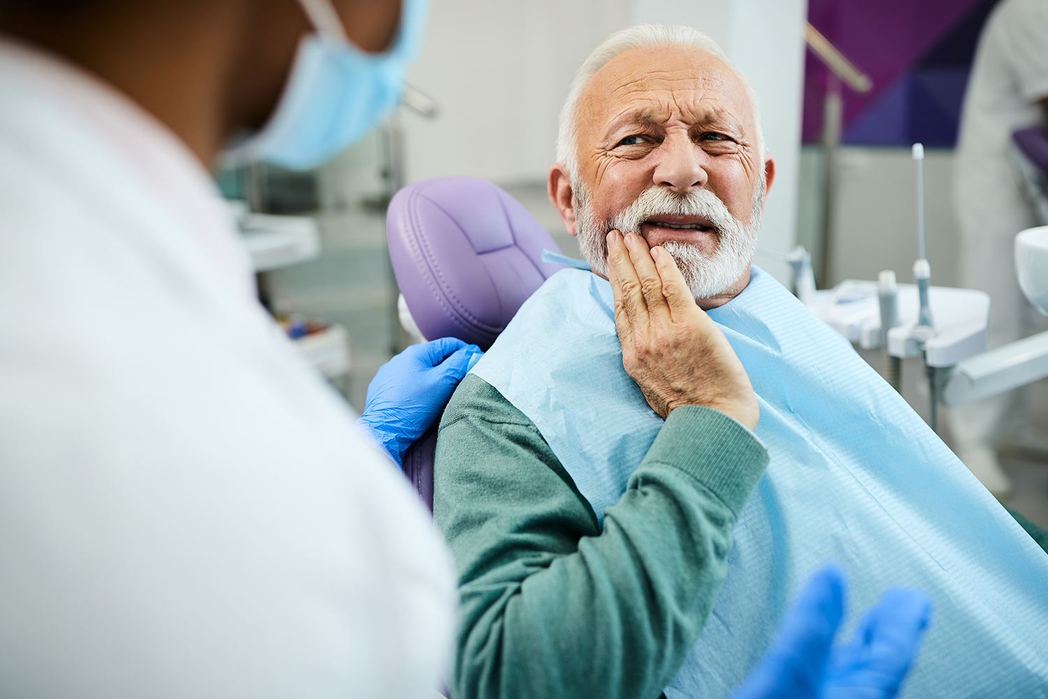 An elderly man is sitting in a dental chair holding his mouth in pain.