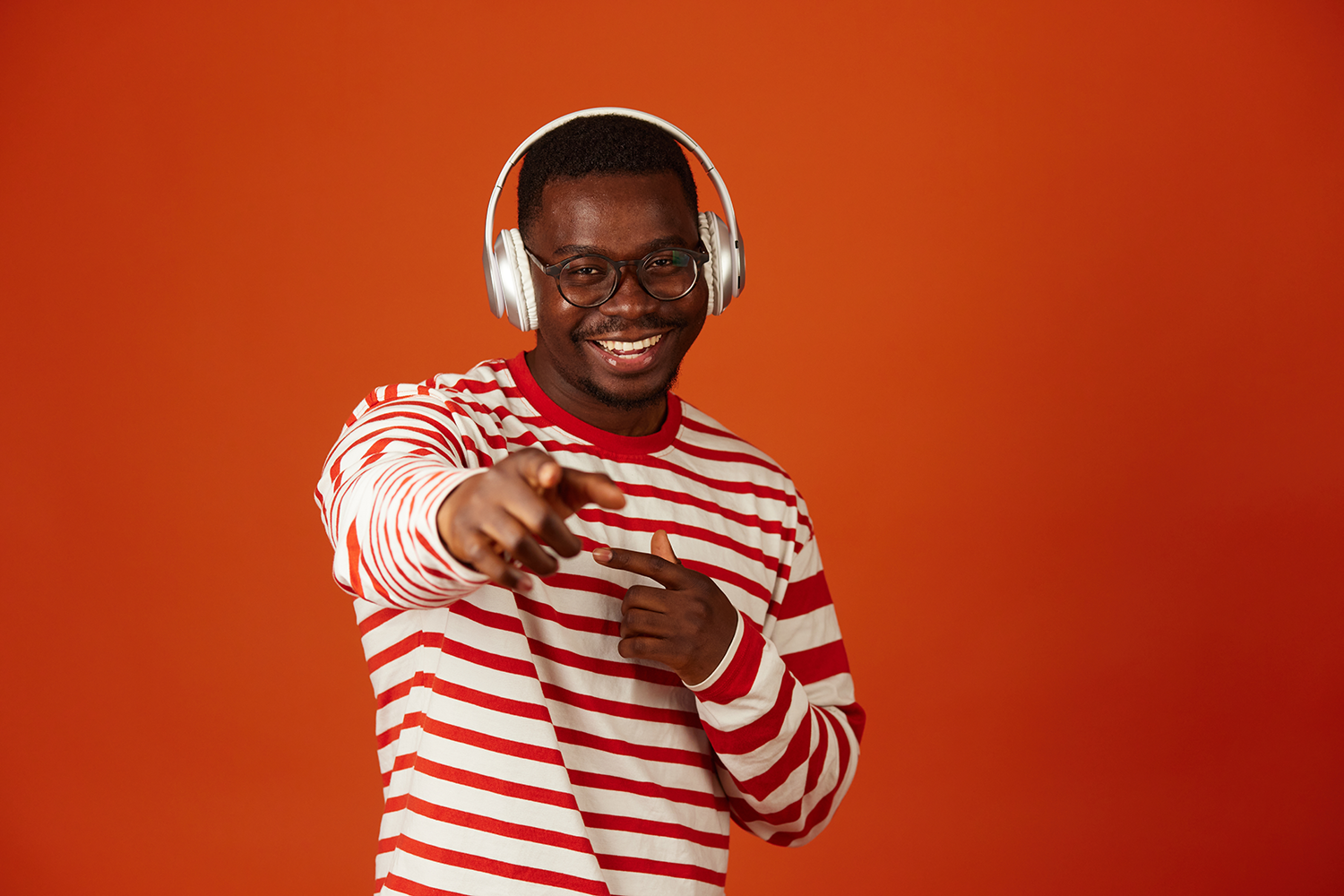 A man wearing headphones and a striped sweater is pointing at the camera.