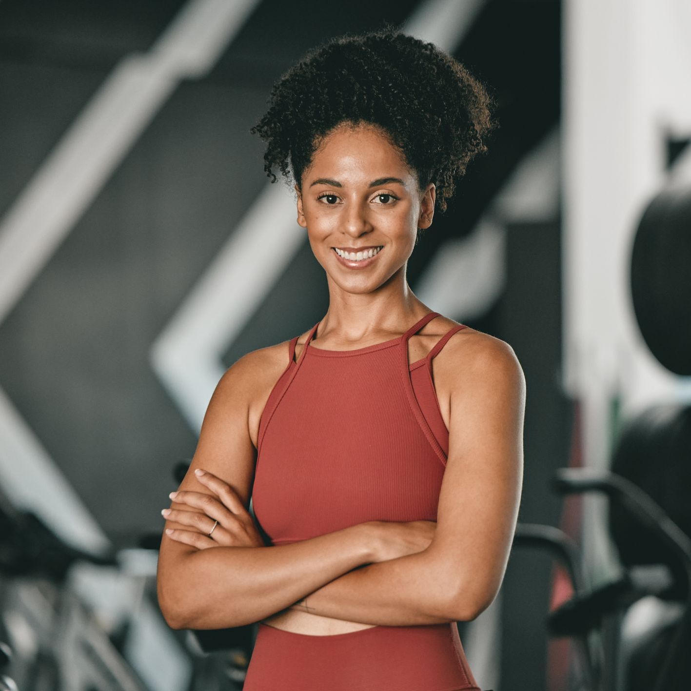 A woman is standing in a gym with her arms crossed and smiling.
