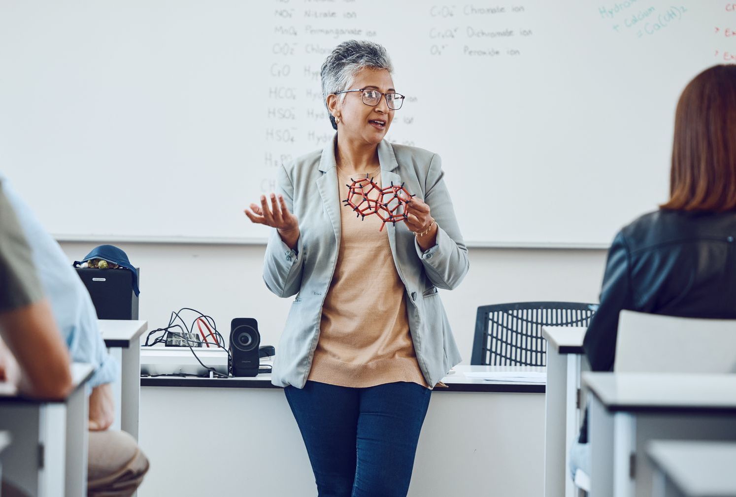 A woman is standing in front of a classroom holding a model of a molecule