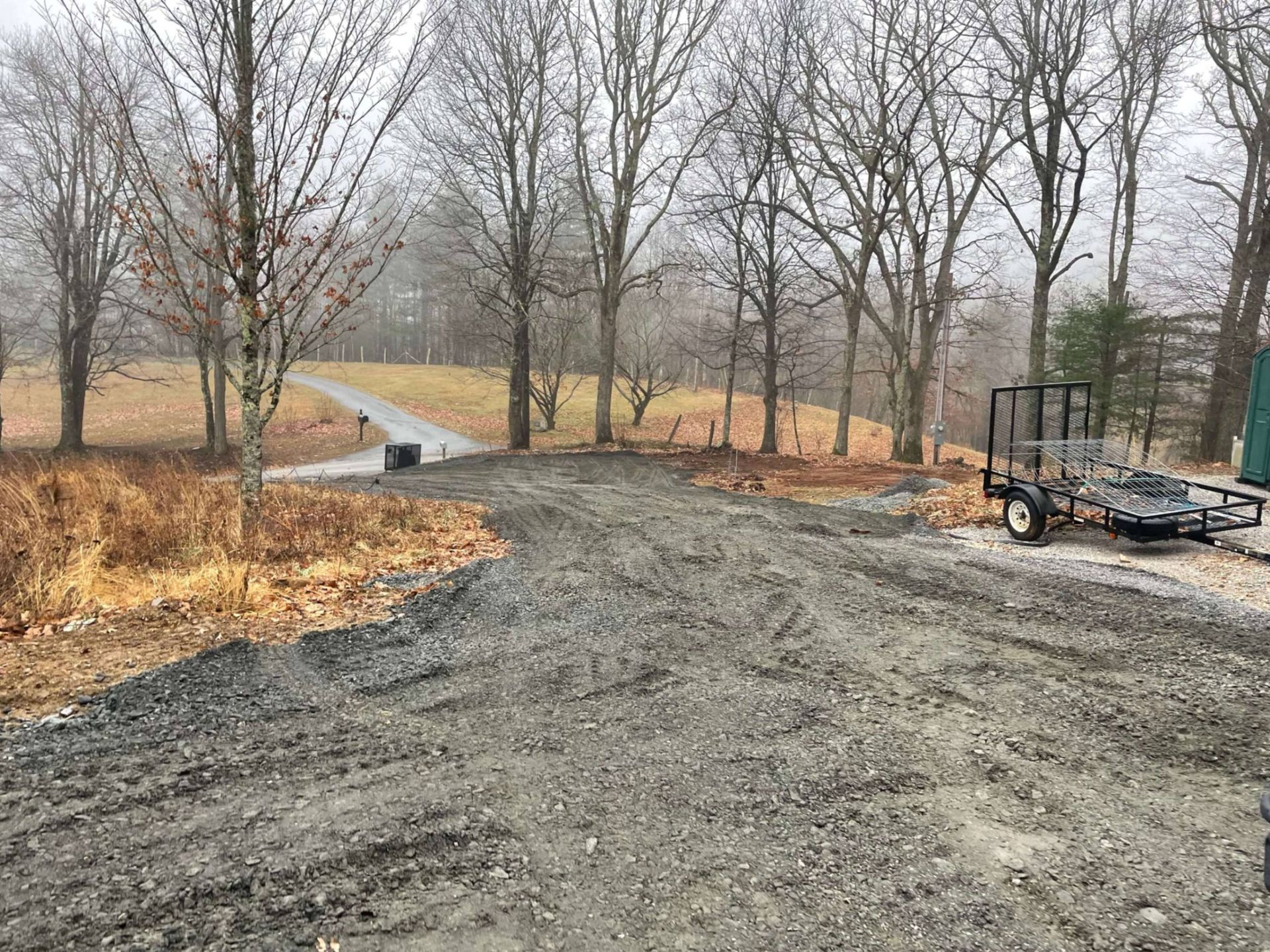 A gravel driveway with a trailer, leading to a path through bare trees on a cloudy day.