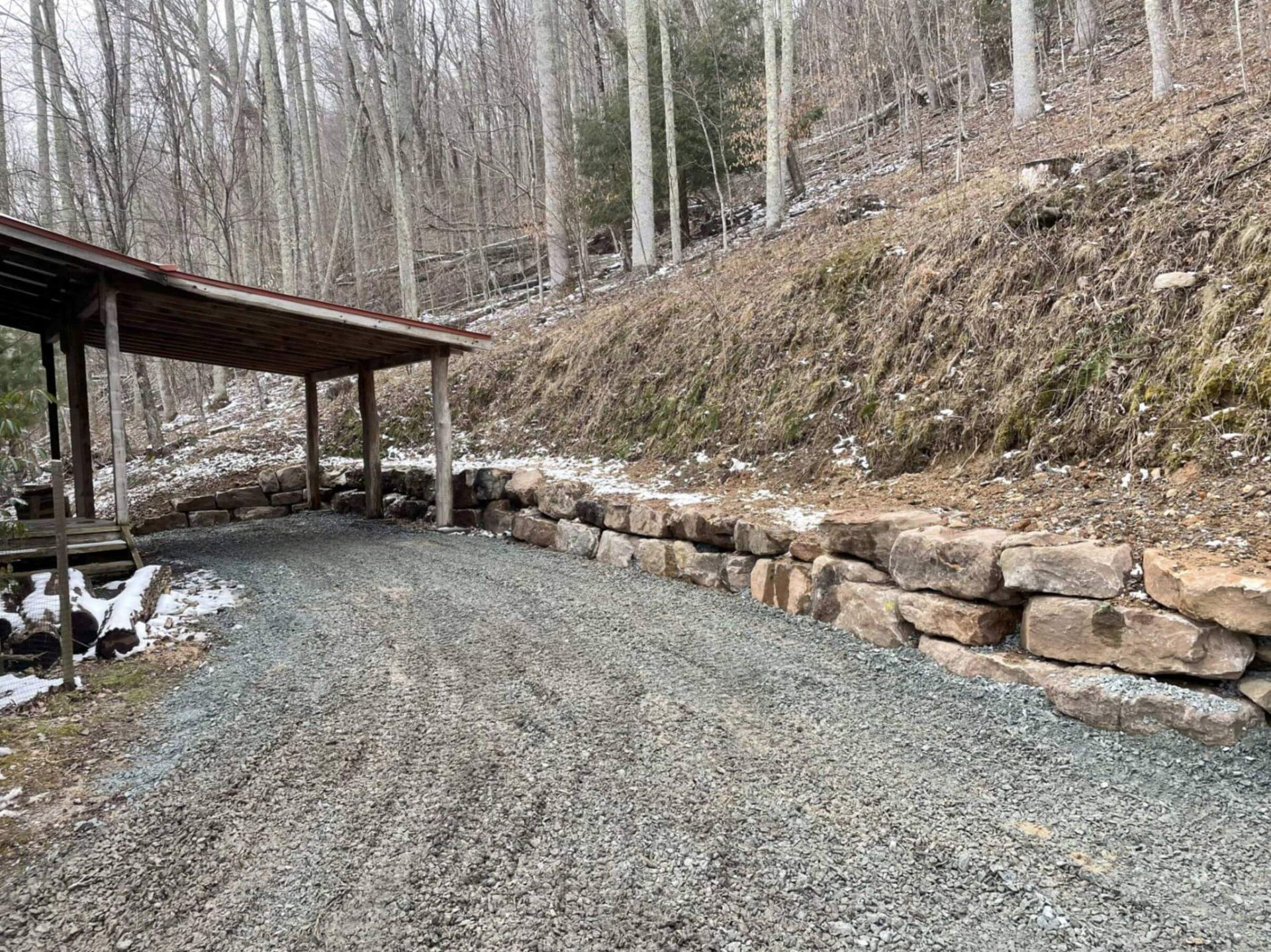 Gravel driveway with a stone retaining wall, leading to a wooden shelter in a wooded area.