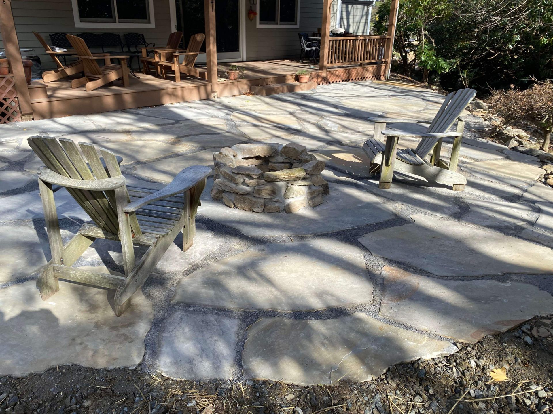 Two wooden Adirondack chairs face a stone fire pit on a stone patio in a backyard.