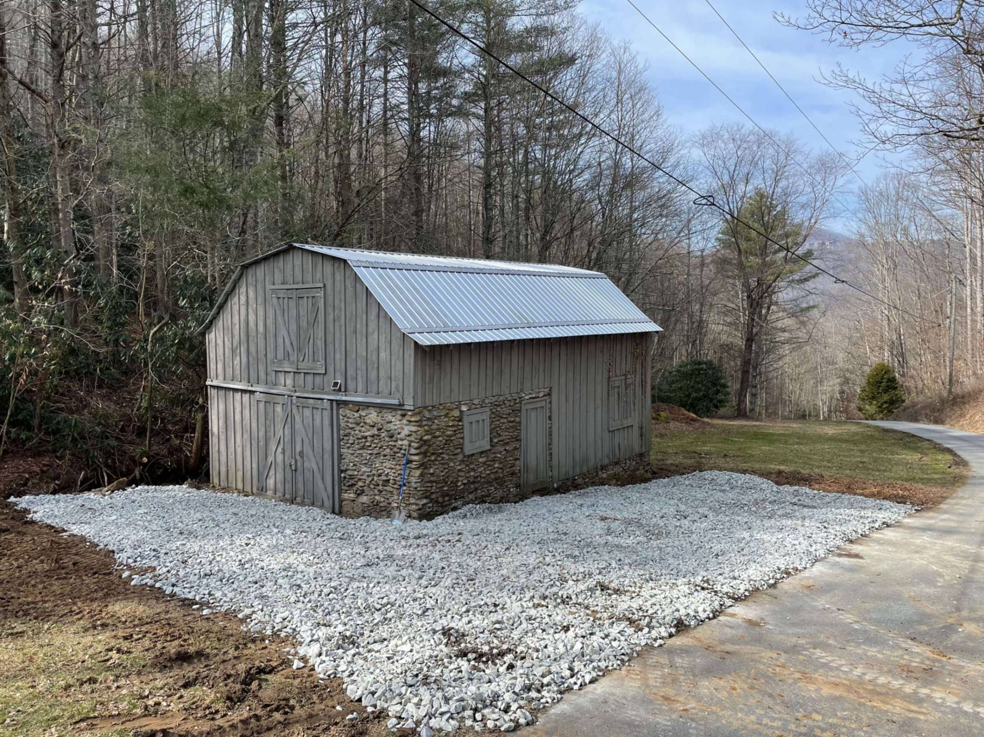 Weathered gray barn with stone foundation and gravel driveway, next to a wooded road.
