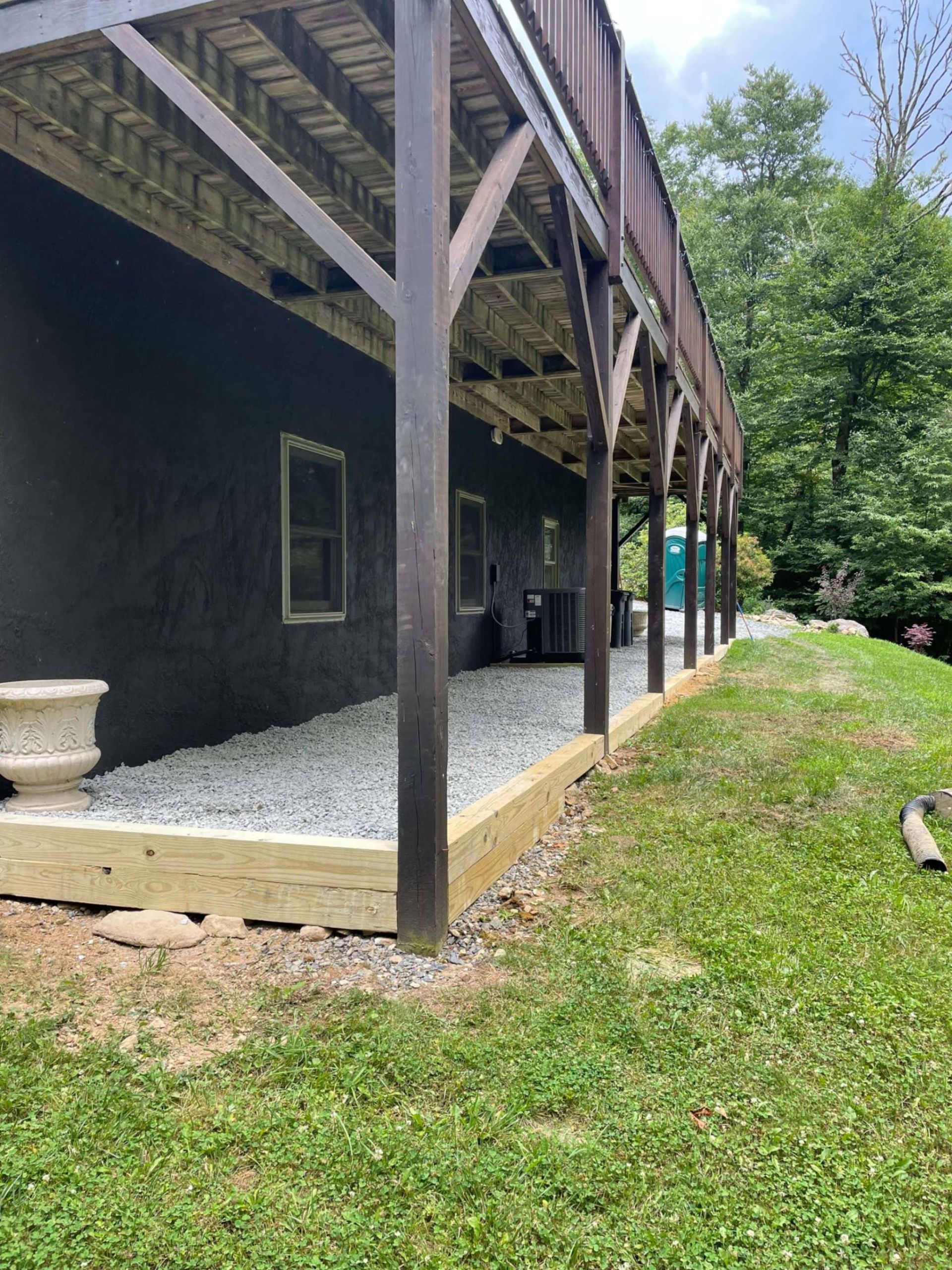 Side view of a dark building with a wooden deck above, a gravel pathway below, and green grass.