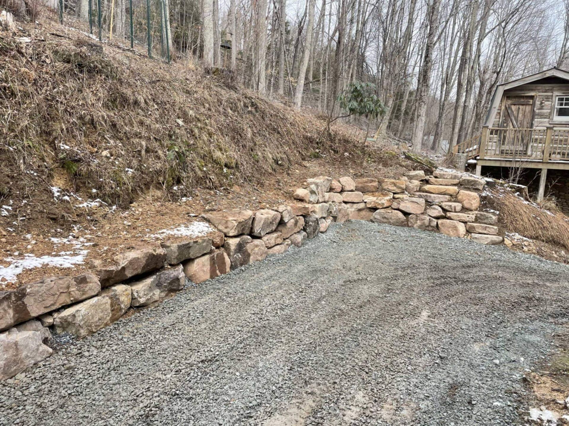 Stone retaining wall beside a gravel pile on a hillside, with a cabin visible in the background.