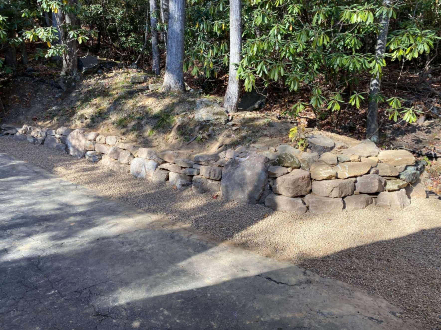 Stone retaining wall along a gravel driveway in a wooded area with trees and sunlight.