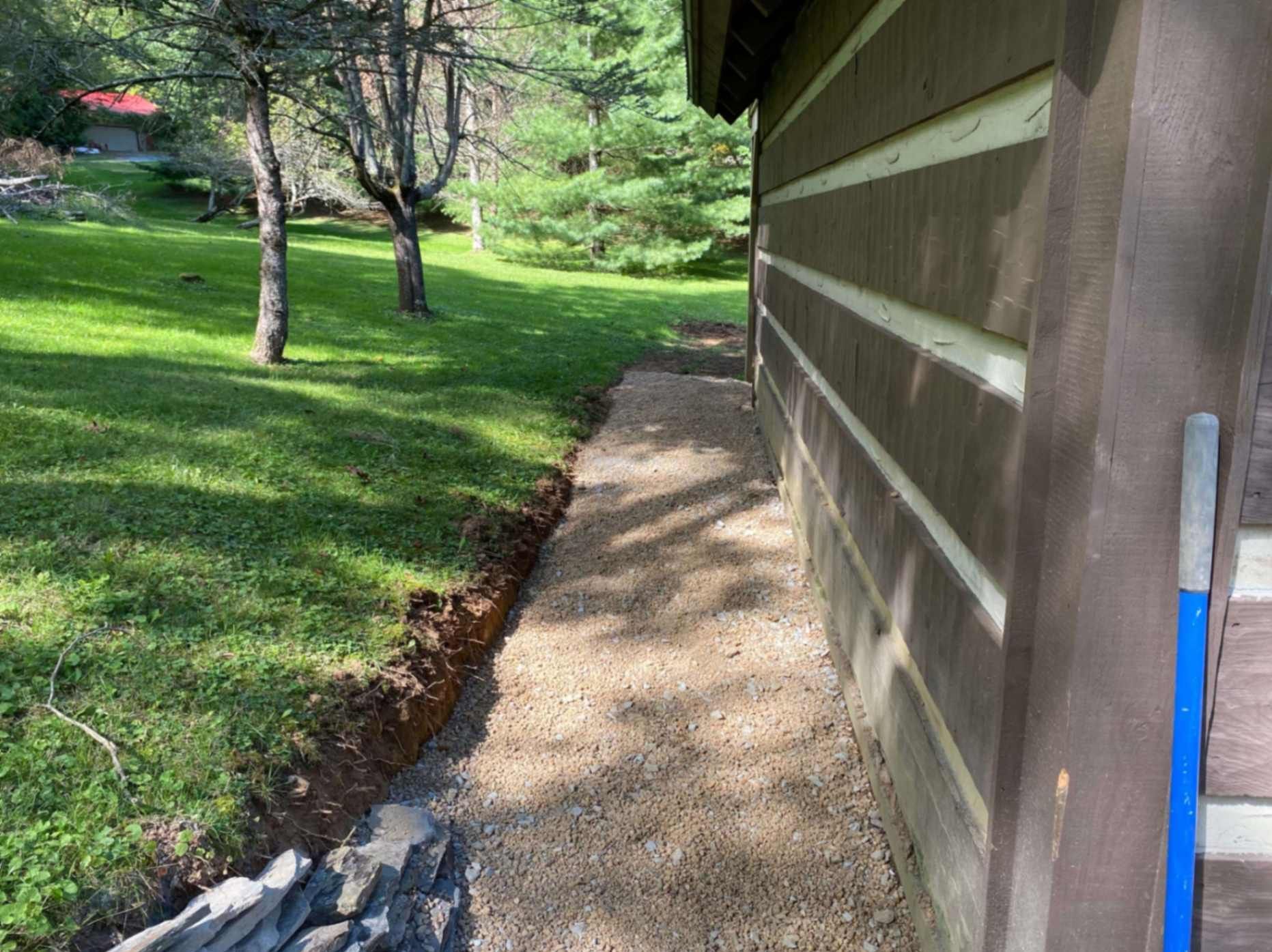 Gravel path next to a brown building with green grass and trees in the background. Blue handle of a tool visible.