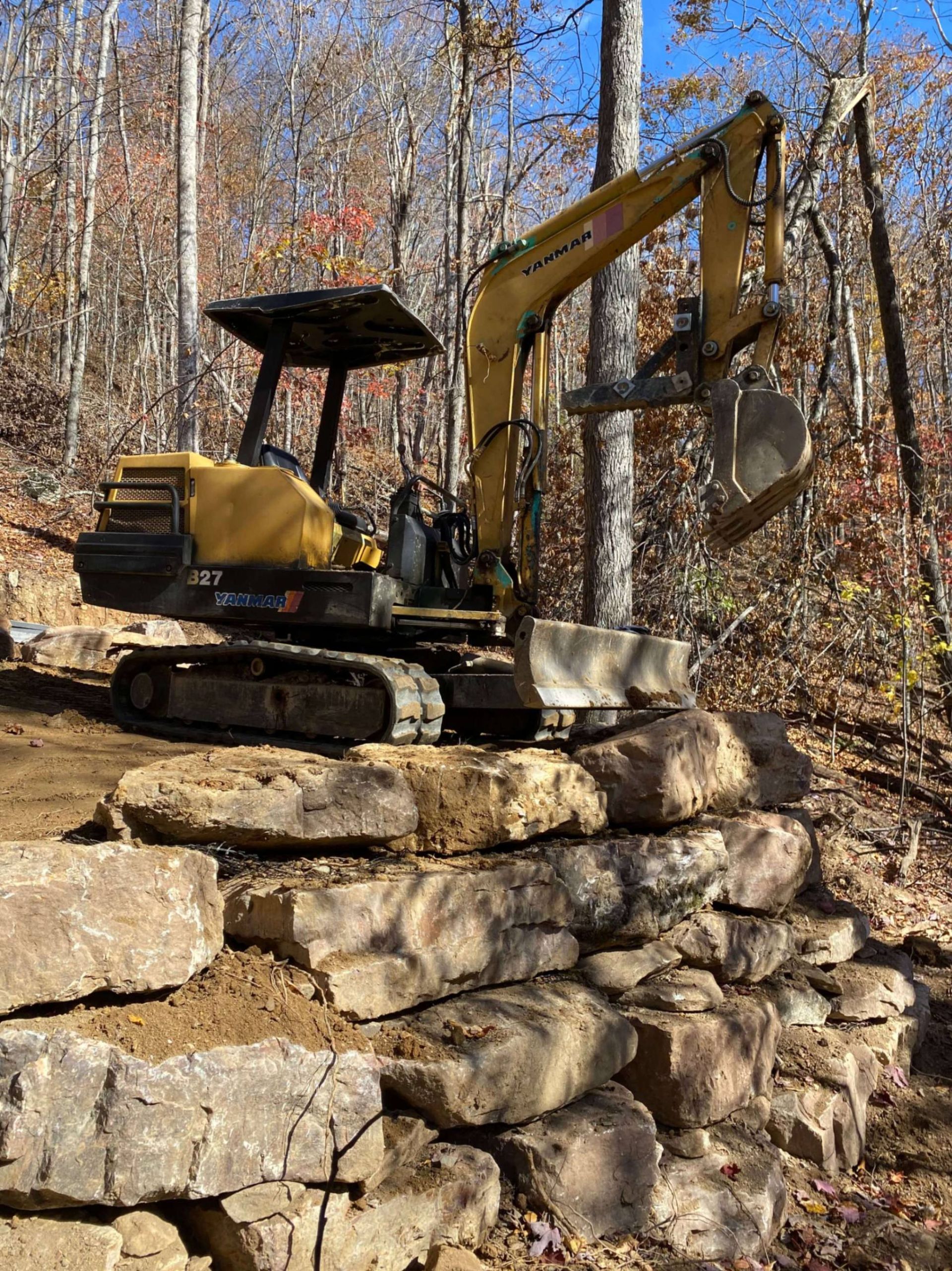 Yellow excavator placing a stone on a rock retaining wall in a wooded area.