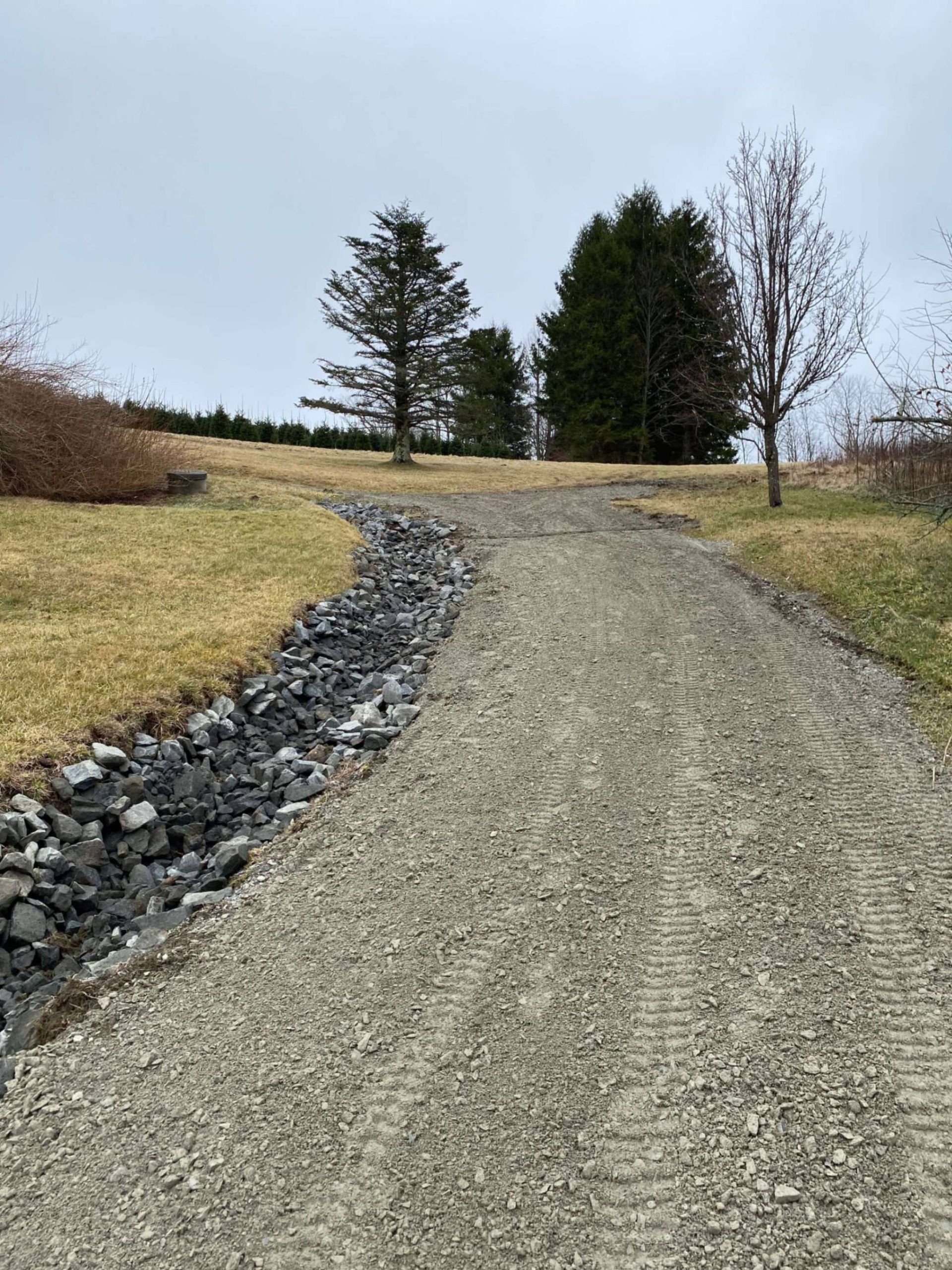 Gravel driveway leading up a hill, with a dark rock-lined drainage ditch on the left, and trees in the background.