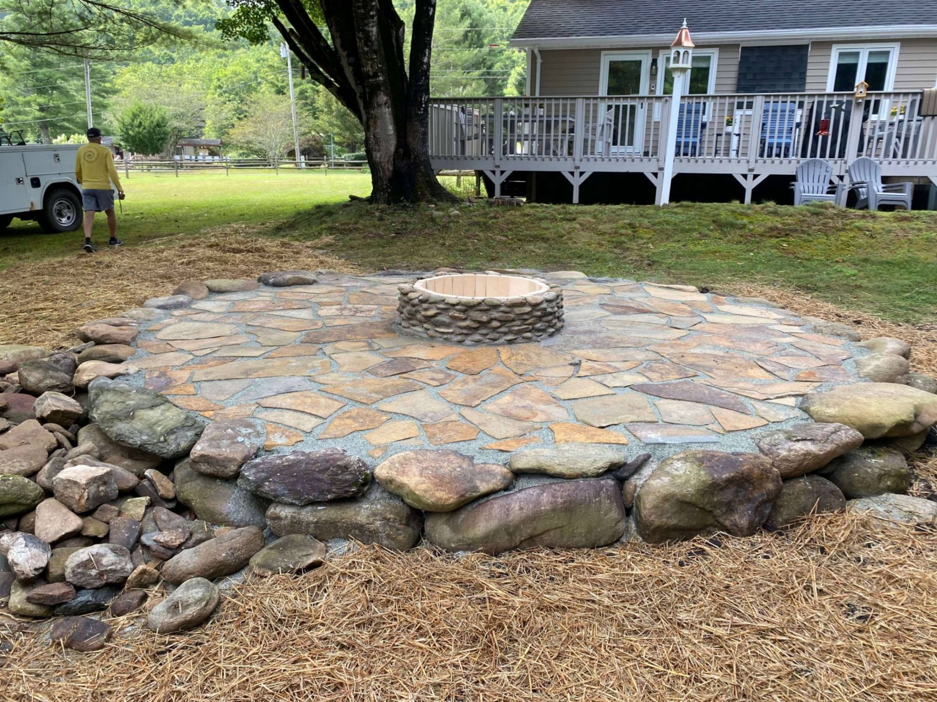 Stone fire pit with rock border in a yard; a man walks near a house with a deck.