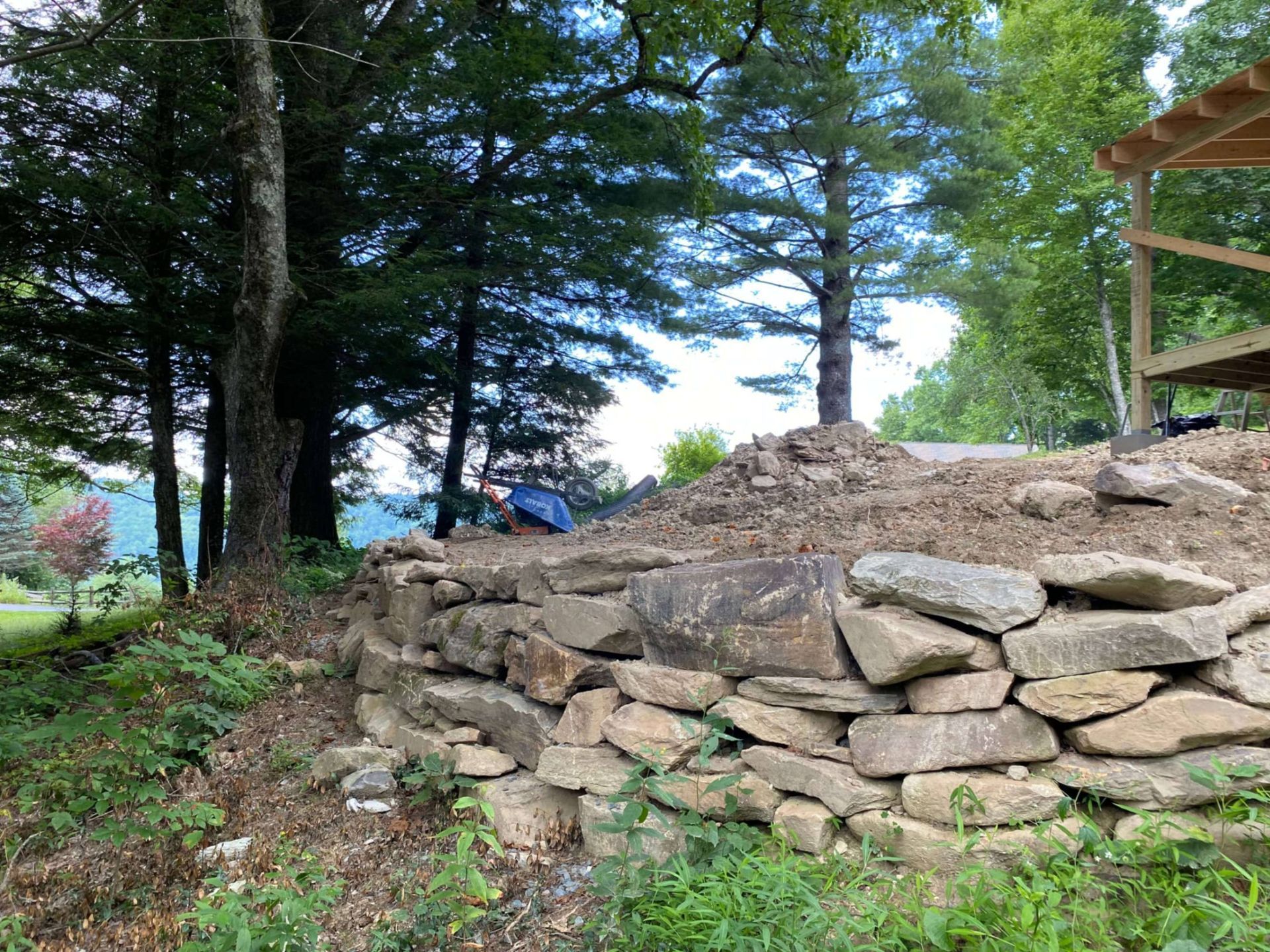 A dry-stack stone retaining wall holds back a mound of earth beneath mature trees and a wooden structure.