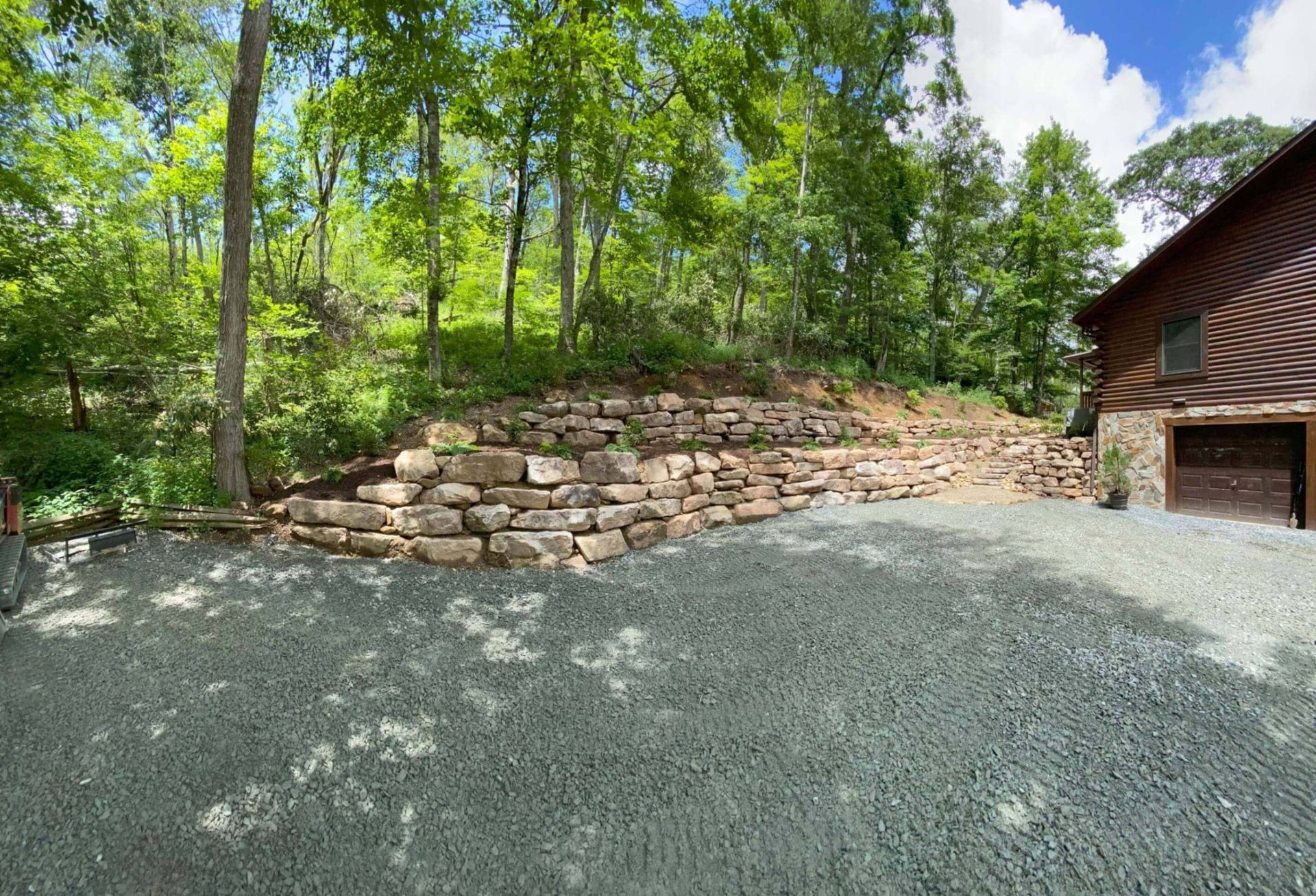 Gravel driveway with stone retaining wall, trees, and brown house with garage door.