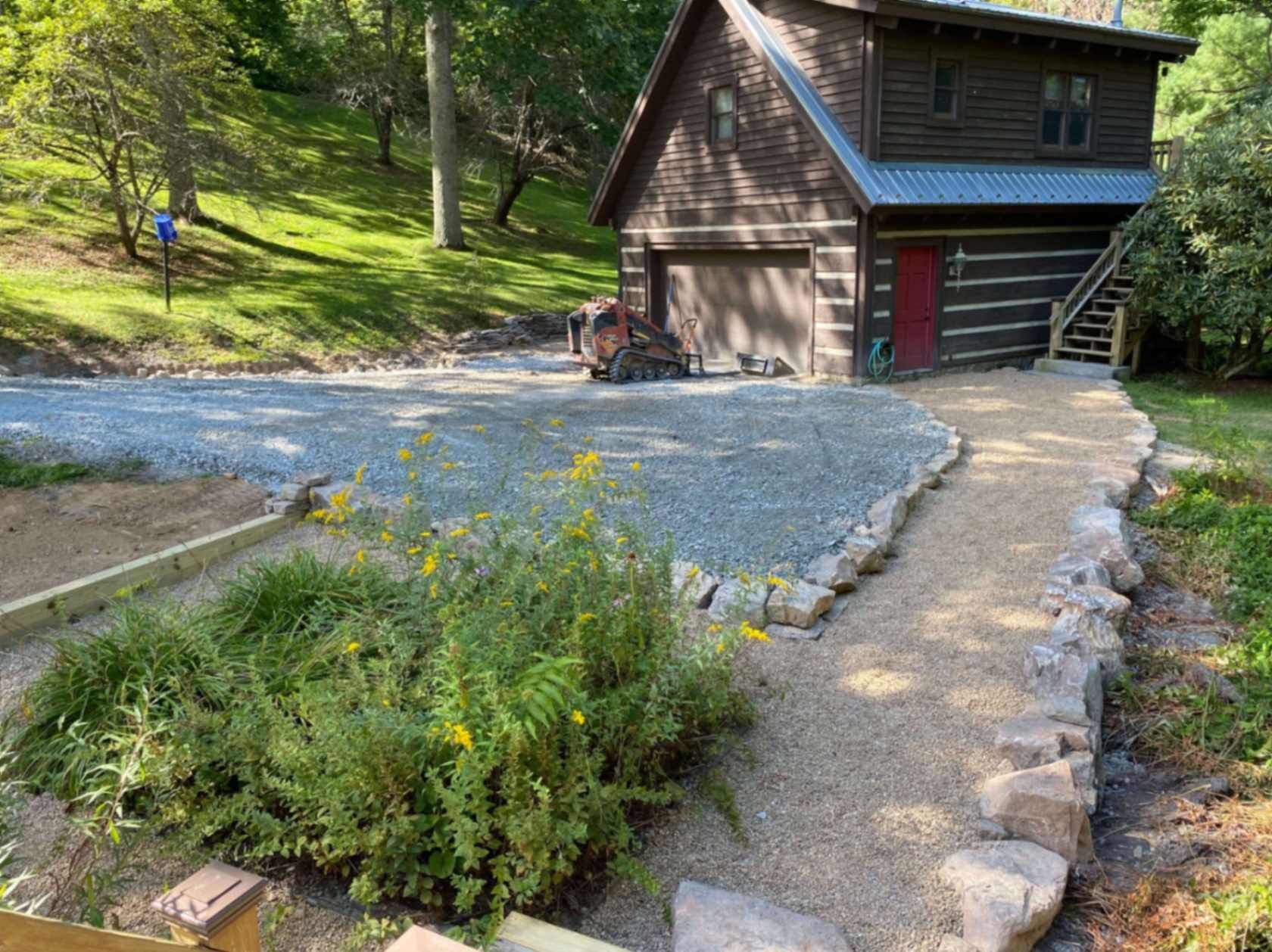 Gravel driveway and pathway leading to a two-story log cabin with a garage door and red door. Green grass and trees surround.