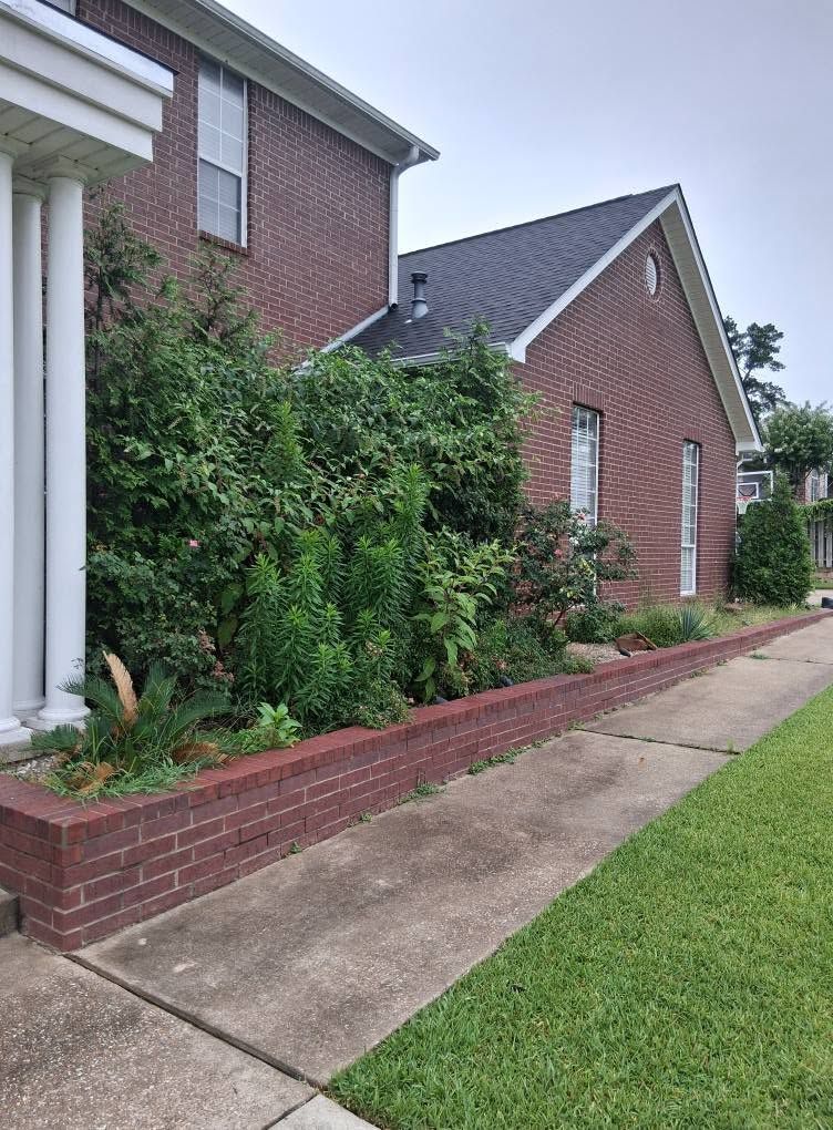 Brick house exterior with a brick planter, lush green shrubs, and a concrete sidewalk along the lawn