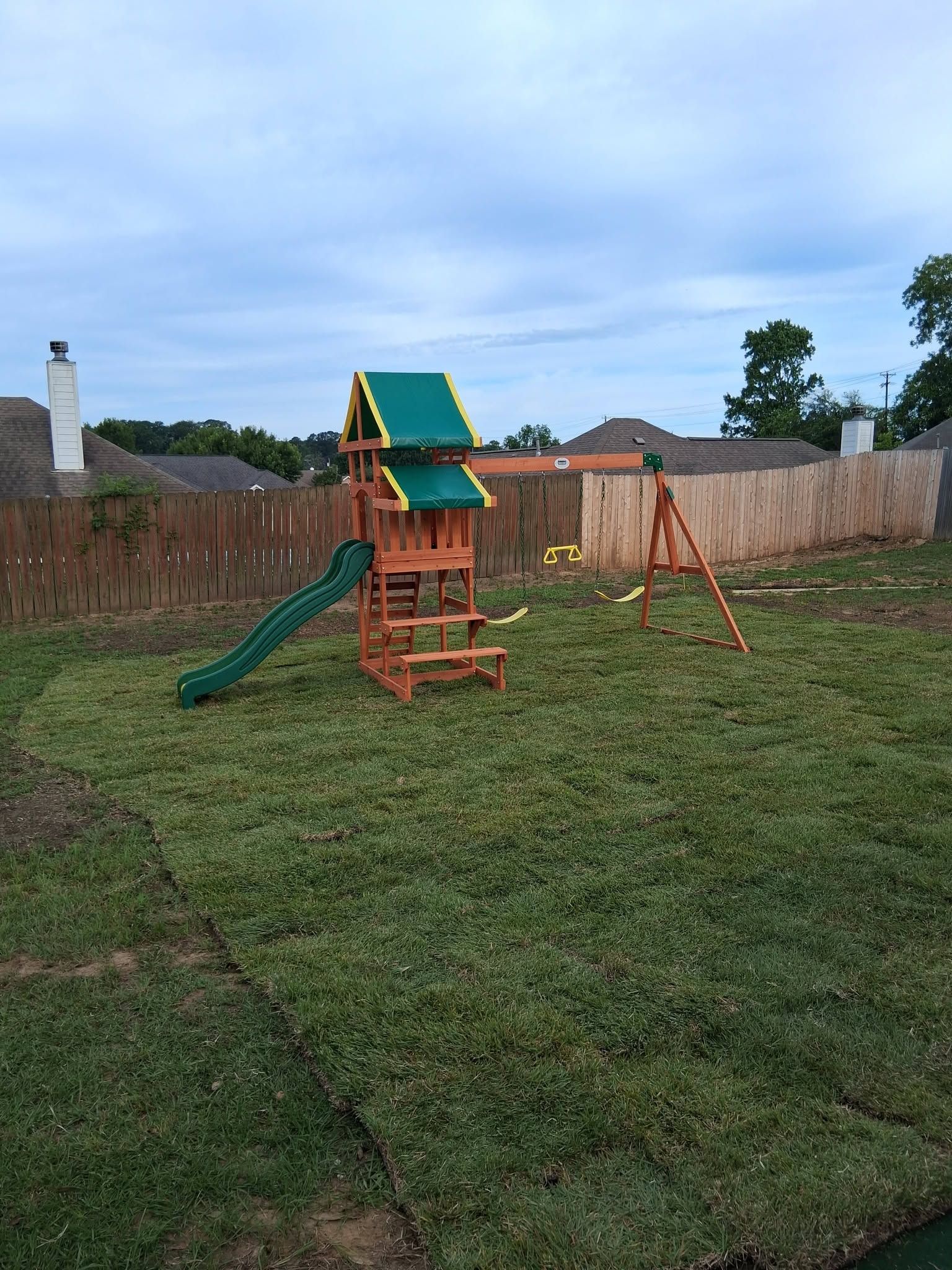 Backyard playground with green slide, swing set, and wooden play structure on grass under cloudy sky