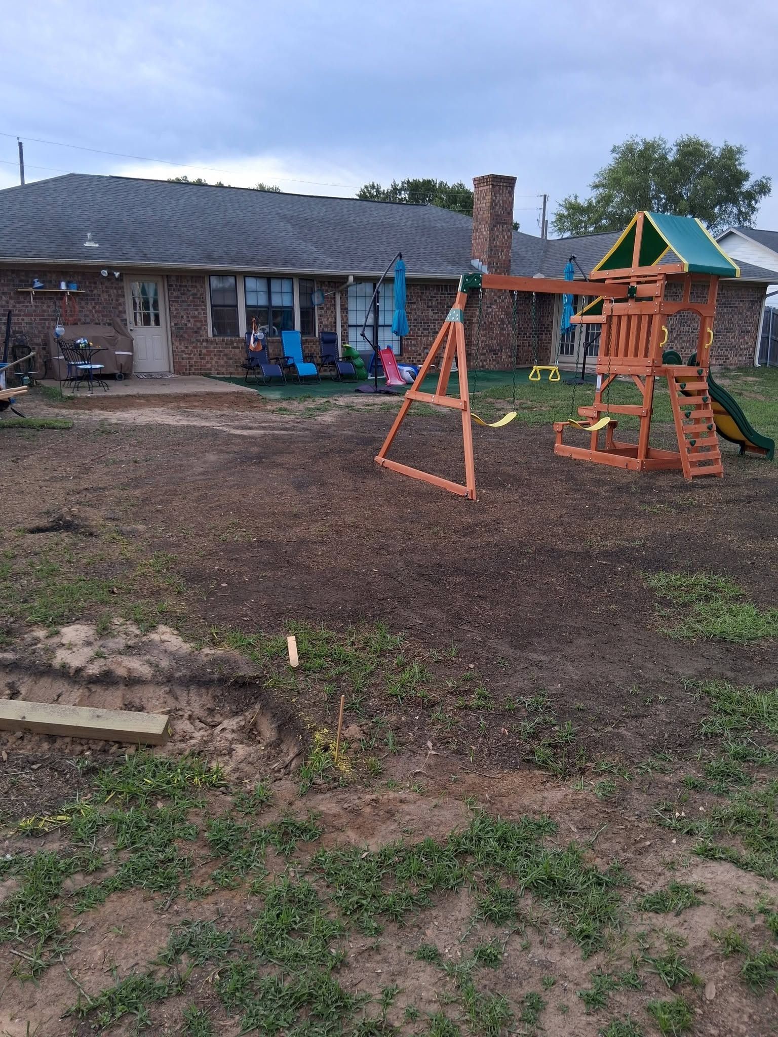 Rural yard with a brick house, muddy ground, and a wooden playset beside an orange swing frame.