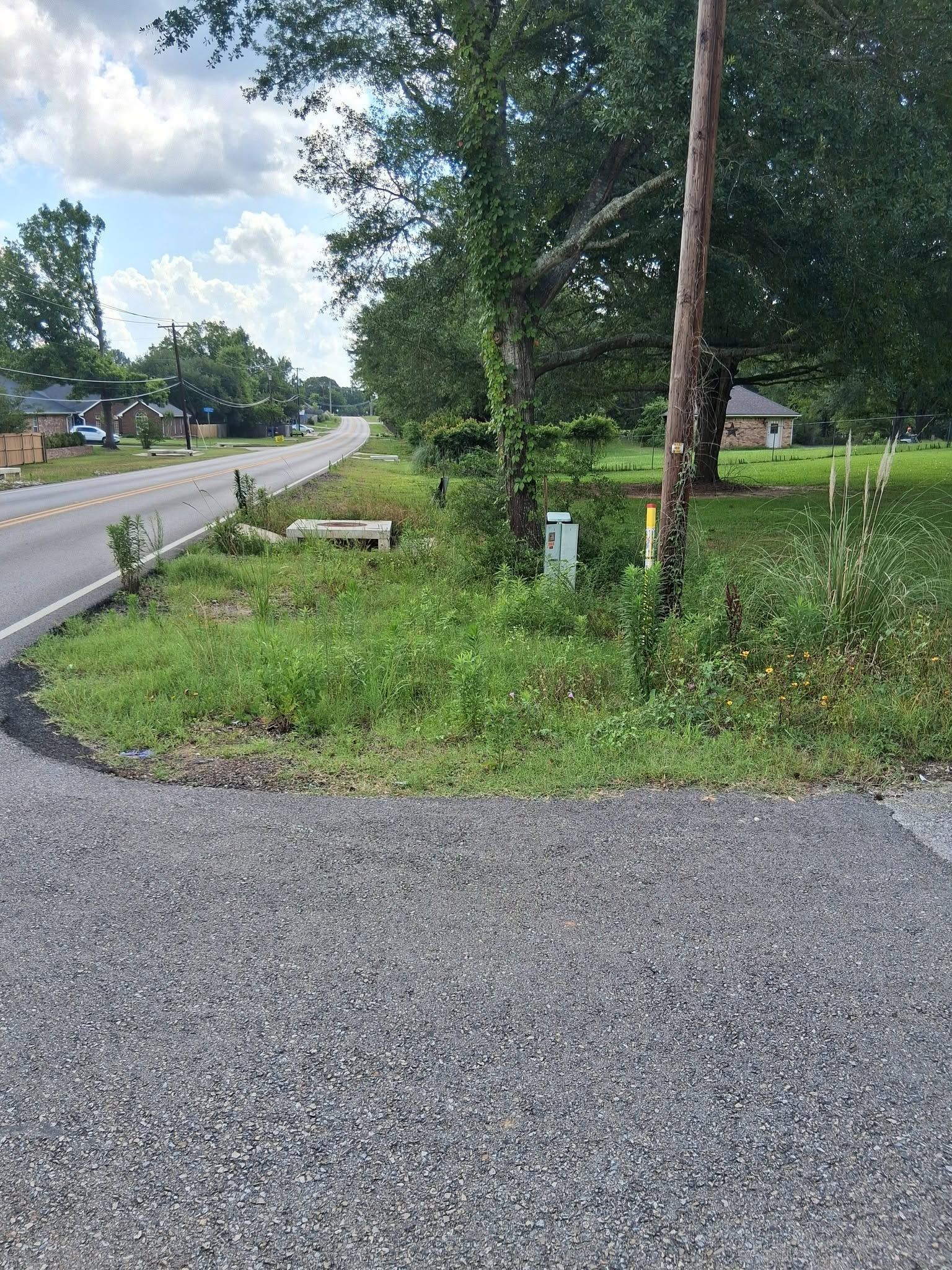 Gravel roadside beside a grassy yard and trees under a cloudy sky, with a paved road on the left.
