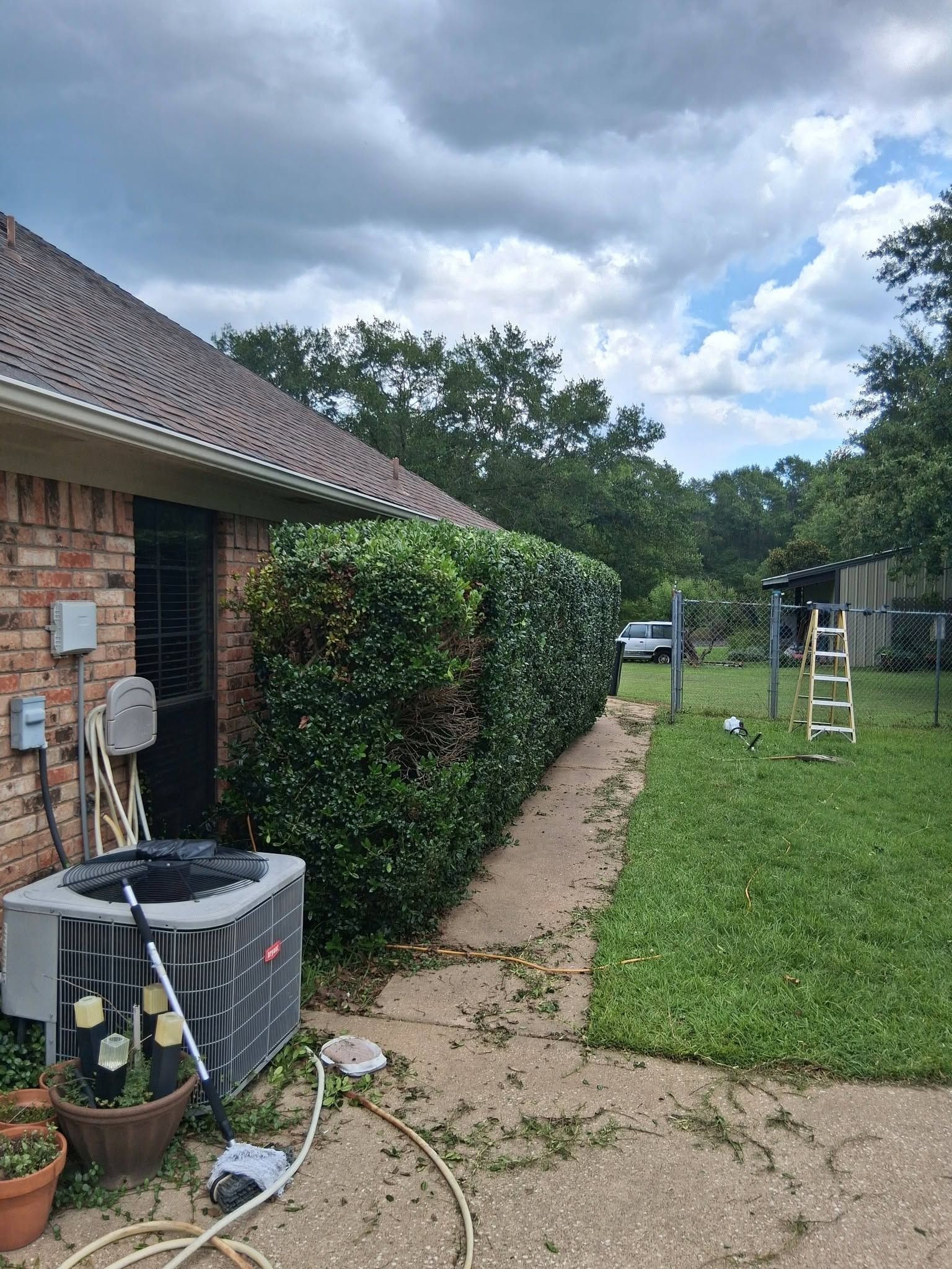 Brick house exterior with air conditioner, trimmed hedge, gravel path, and grassy backyard under cloudy sky