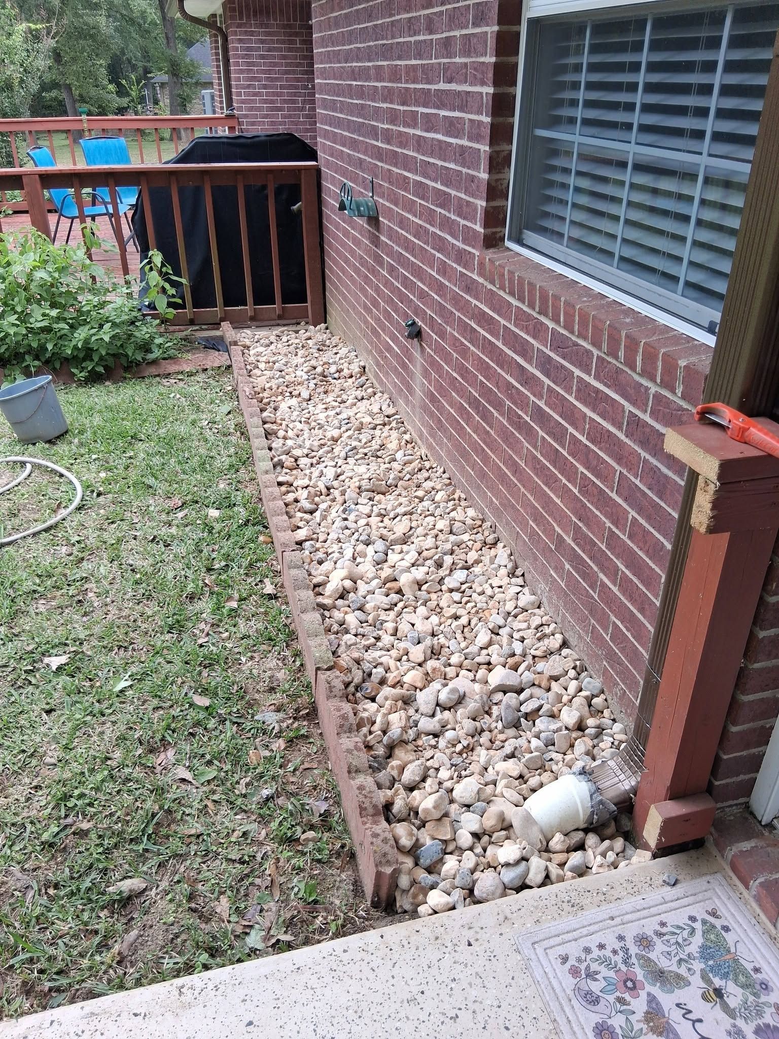 Rock garden strip beside a brick house with window, bordered by grass and a wooden fence, with a pipe near the corner