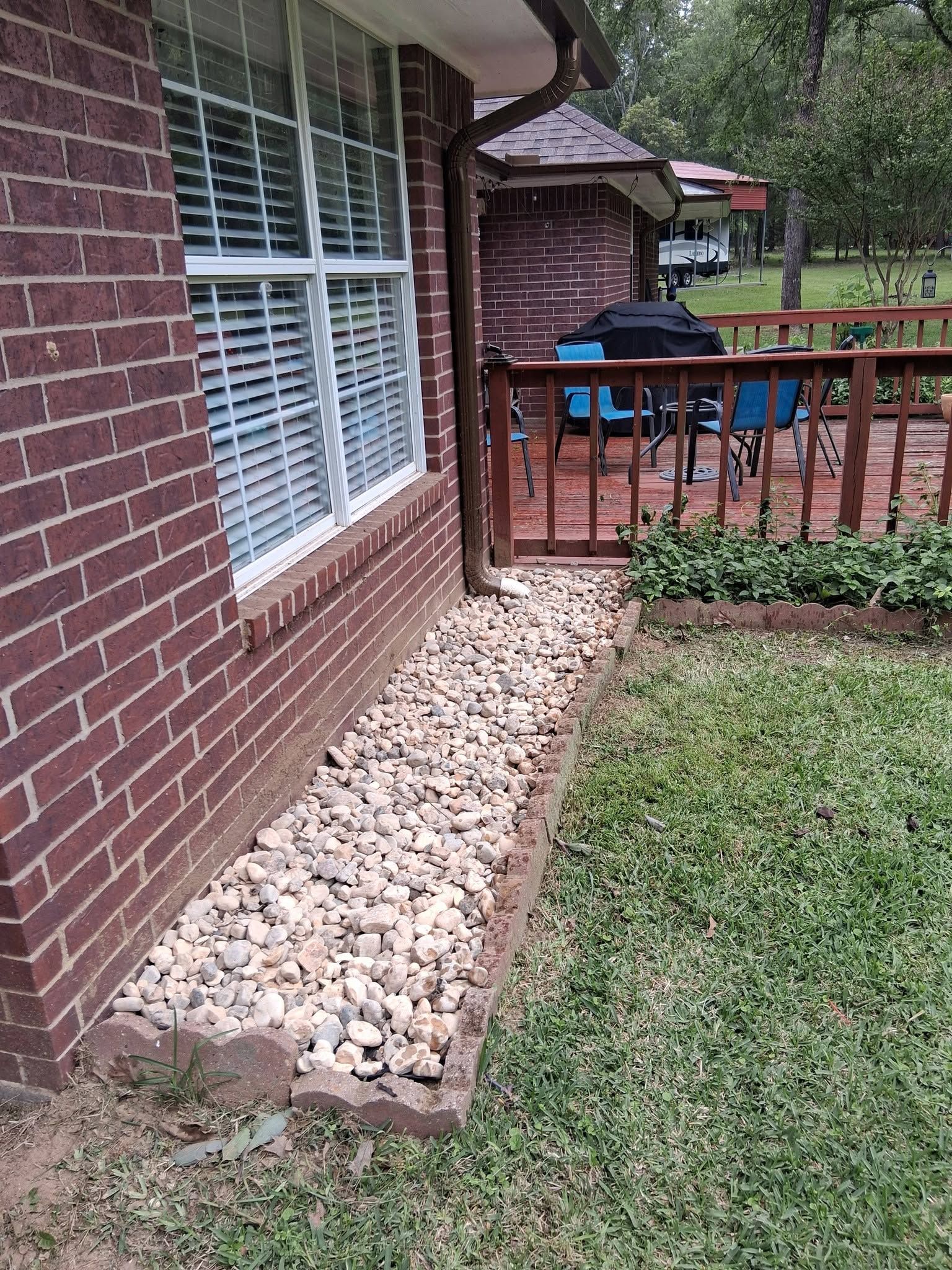 Brick house wall with white-trimmed window beside a rock border and wooden deck railing in a yard