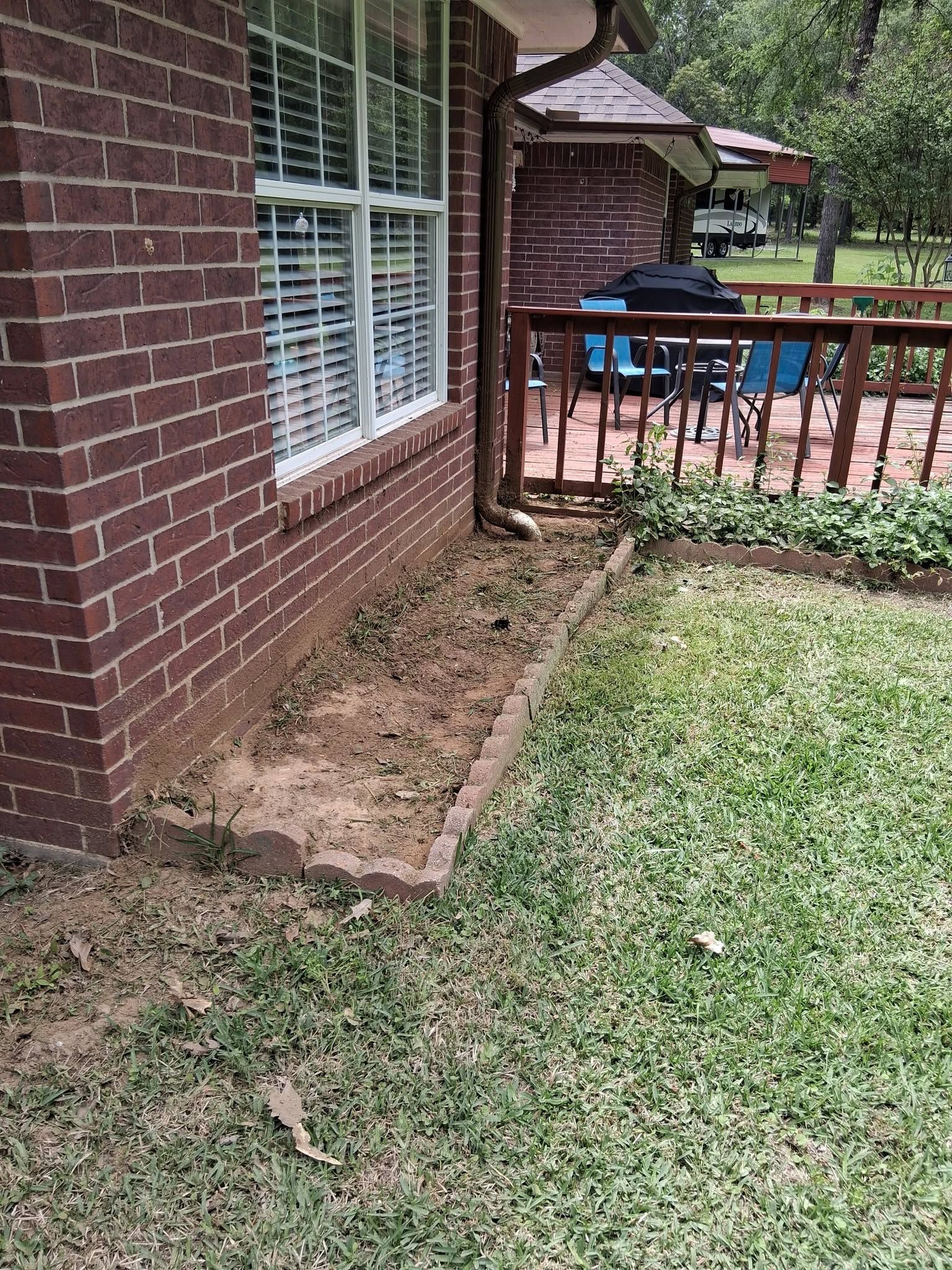 Brick house side yard with a narrow mulched border, grass lawn, and wooden deck railing by a window