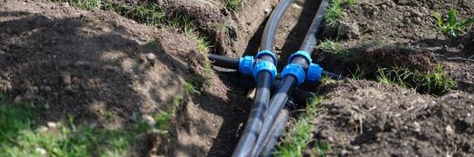 Black irrigation pipes joined by blue connectors in a dirt trench outdoors
