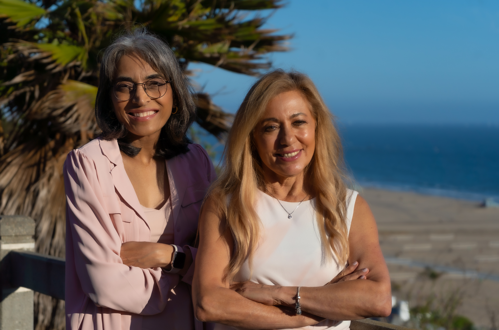 Two women are standing next to each other on a beach with their arms crossed.