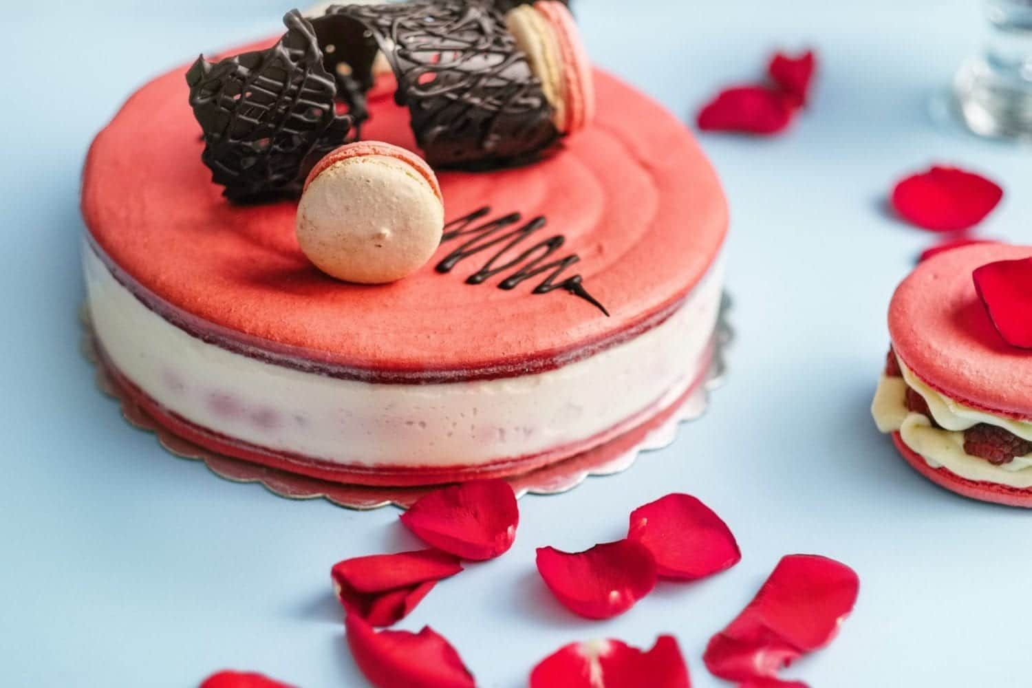 A red and white cake with macarons on top is surrounded by rose petals.