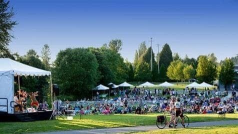 A large group of people are gathered in a park watching a band play on a stage.