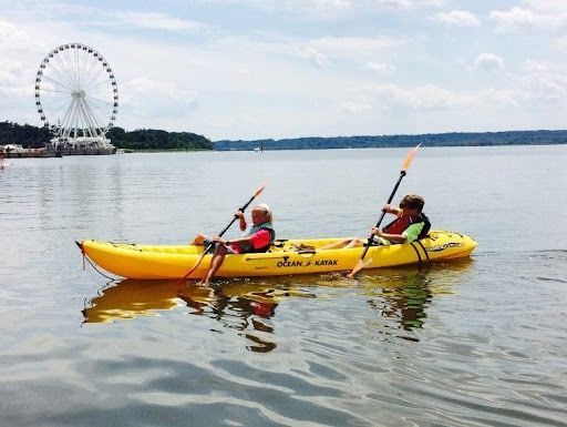 Two people are in a yellow kayak on a lake with a ferris wheel in the background.