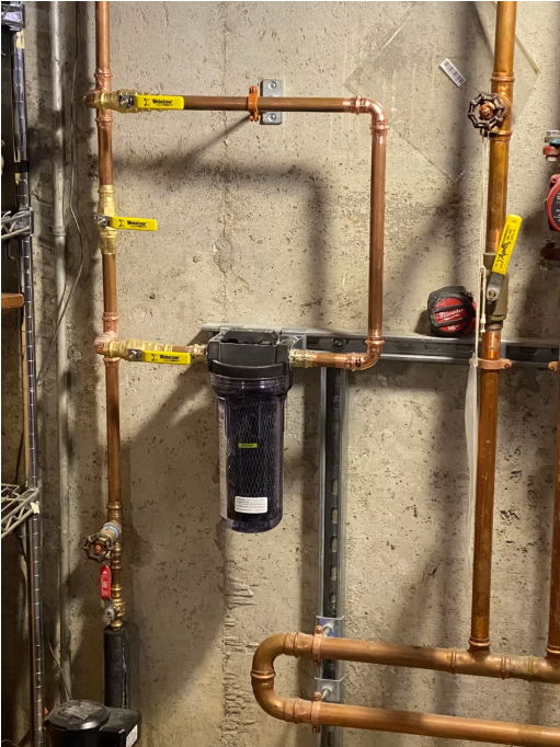 Copper pipes and black water filter installed on a concrete wall with yellow valve handles.