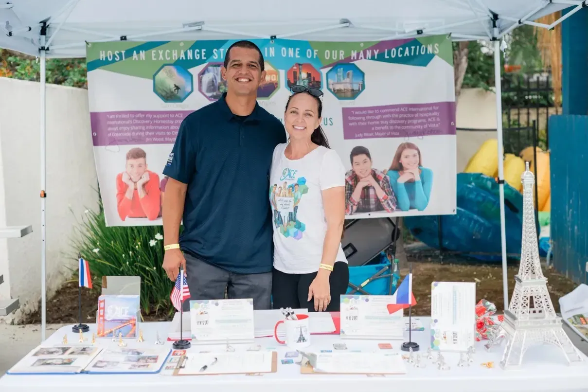 Two people at a booth, smiling. Sign: 