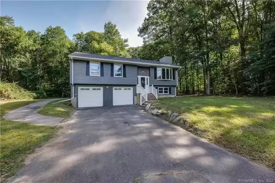 Two-story house with gray siding, two-car garage, and driveway, surrounded by trees and grass under a sunny sky.