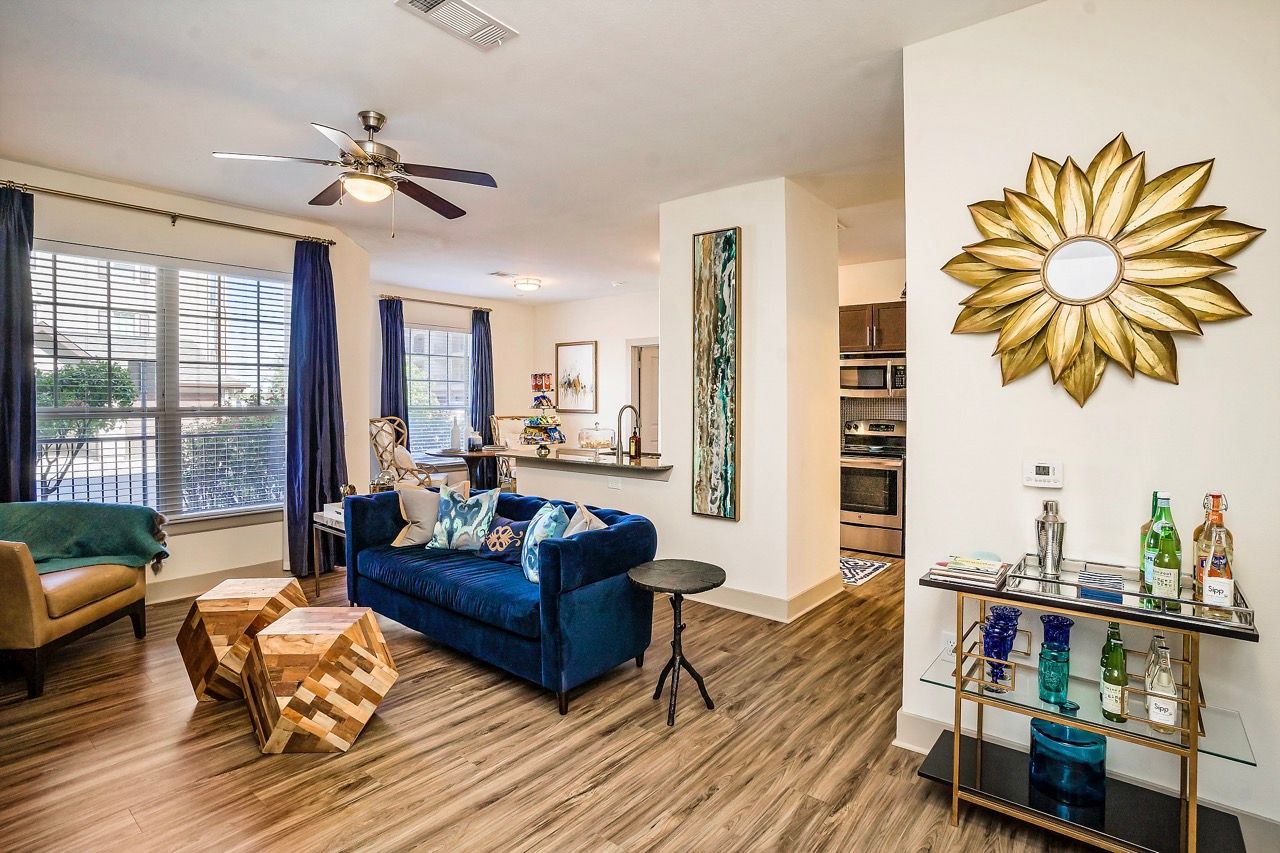 Open-concept living room with blue sofa, wood-style flooring, large windows with dark curtains, and a bar cart.