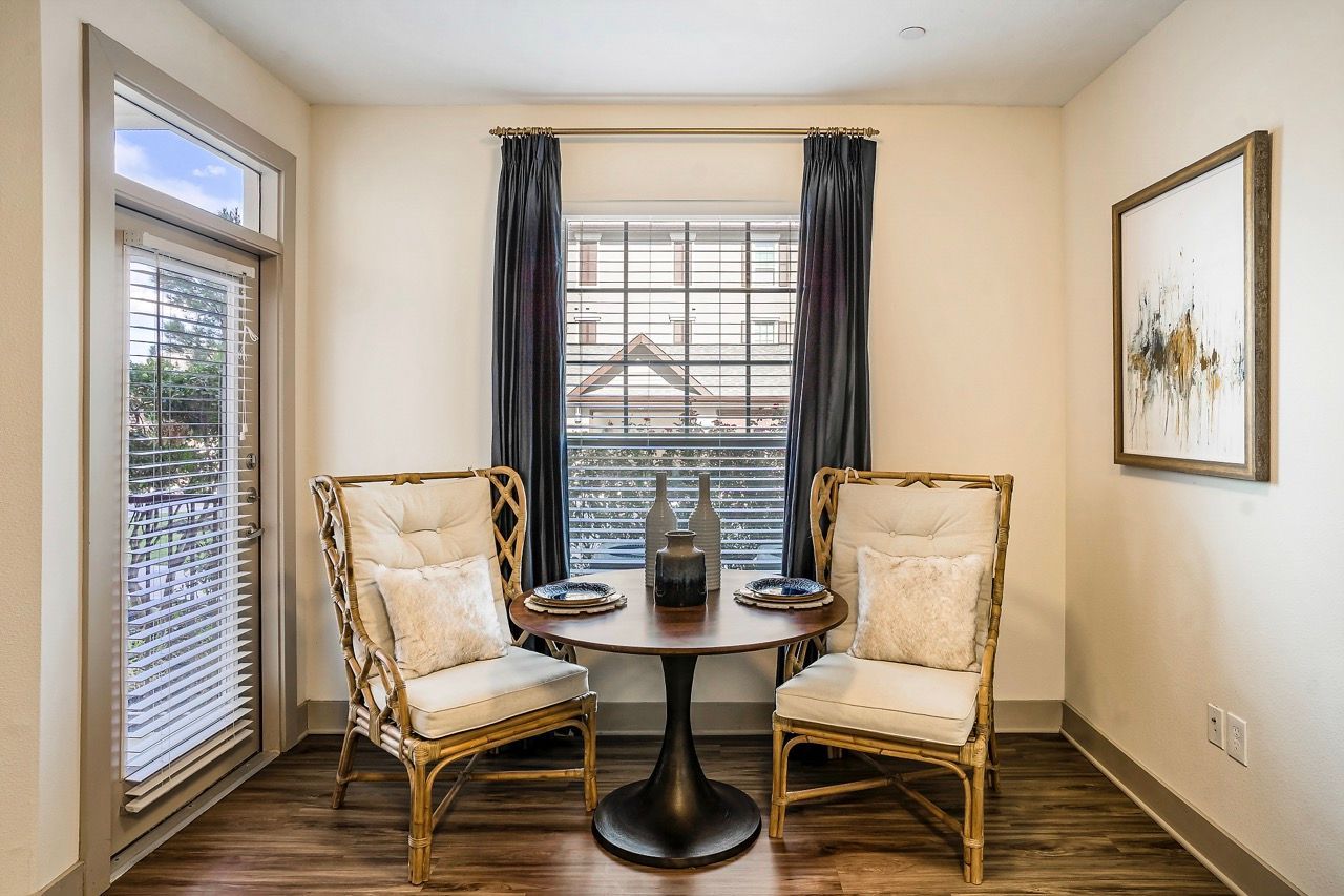 Two cushioned wicker chairs flank a round dining table near a window with blinds and a sliding door.