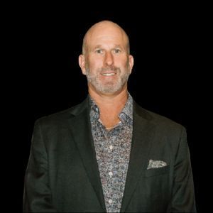Man in a dark blazer and patterned shirt smiling against a white background