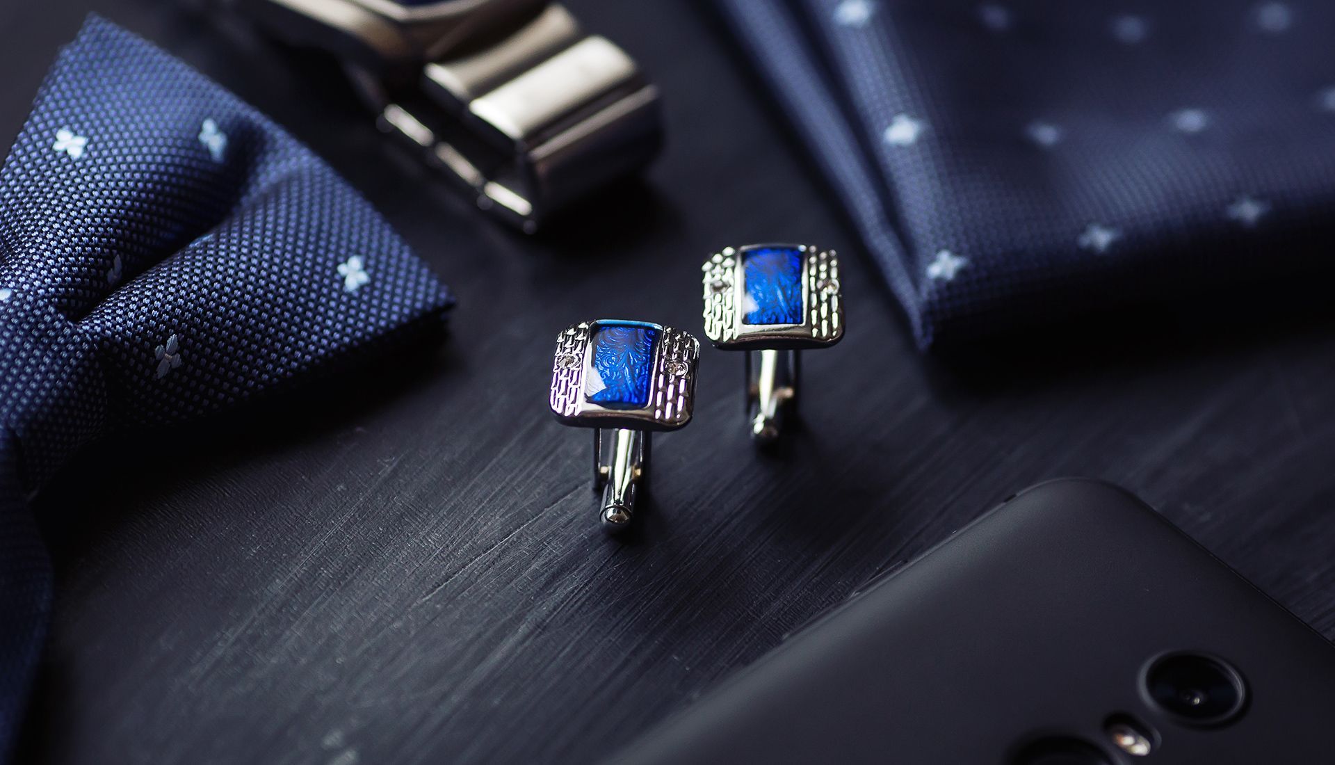 Blue and silver cufflinks with blue gemstone, next to a necktie and handkerchief on a black surface.