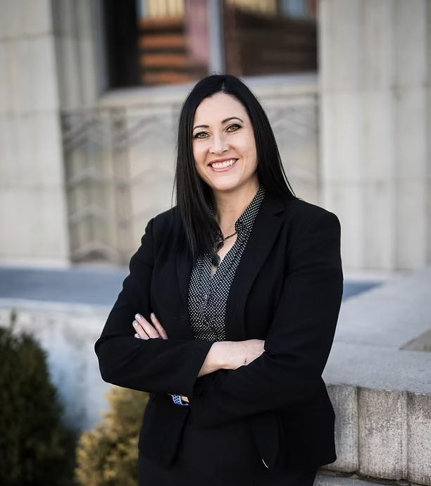A woman in a black suit is smiling with her arms crossed
