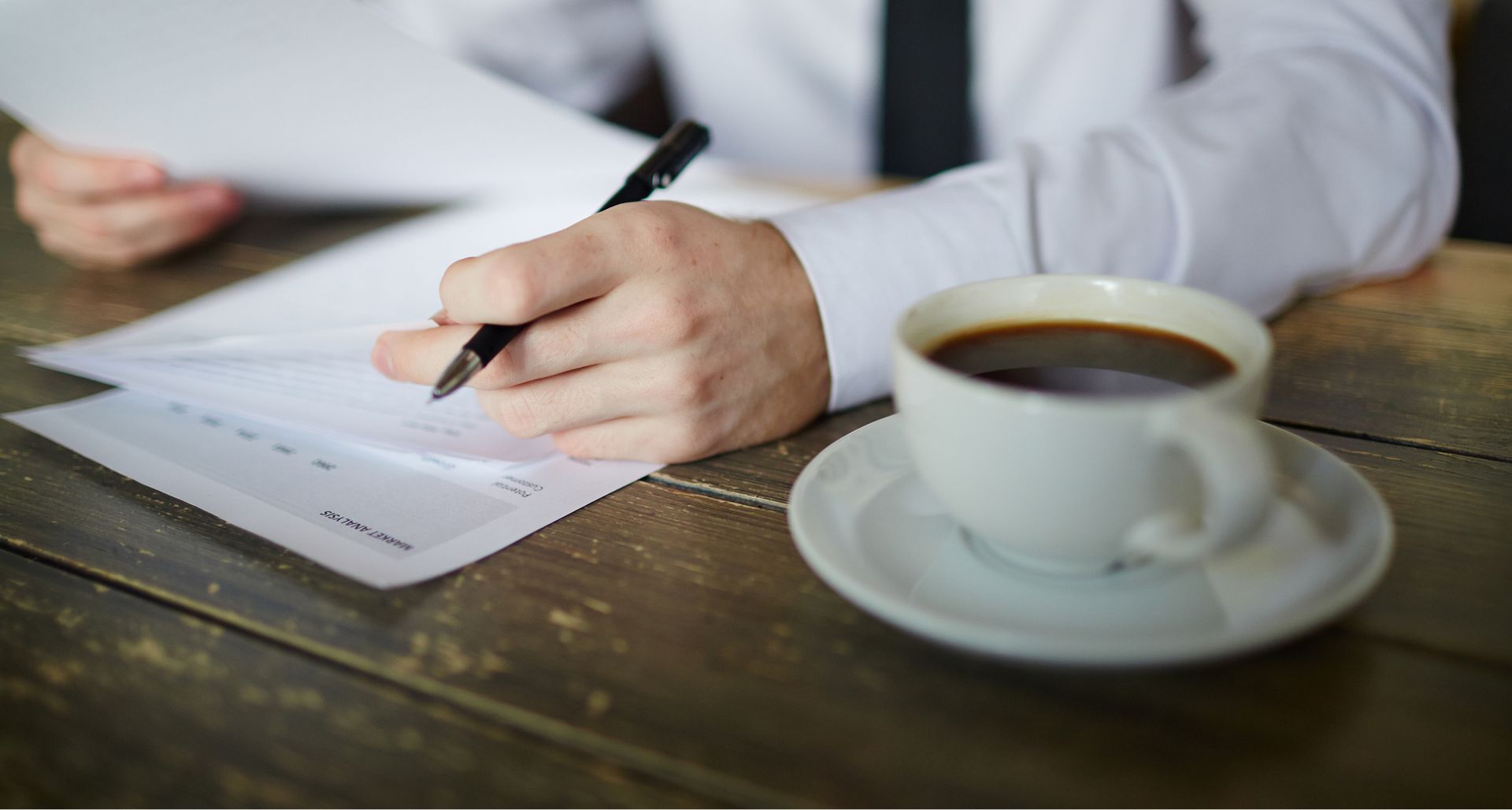A man is writing on a piece of paper next to a cup of coffee.