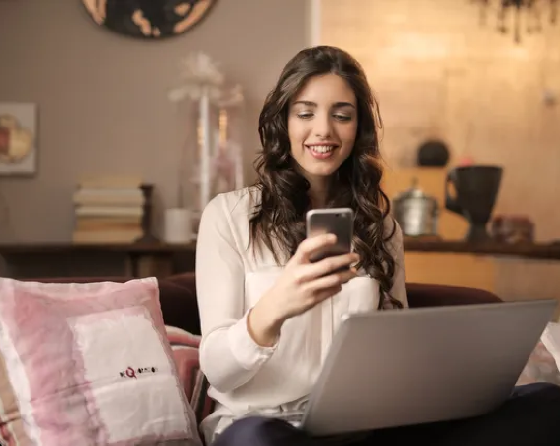 A woman is sitting on a couch using a laptop and a cell phone.