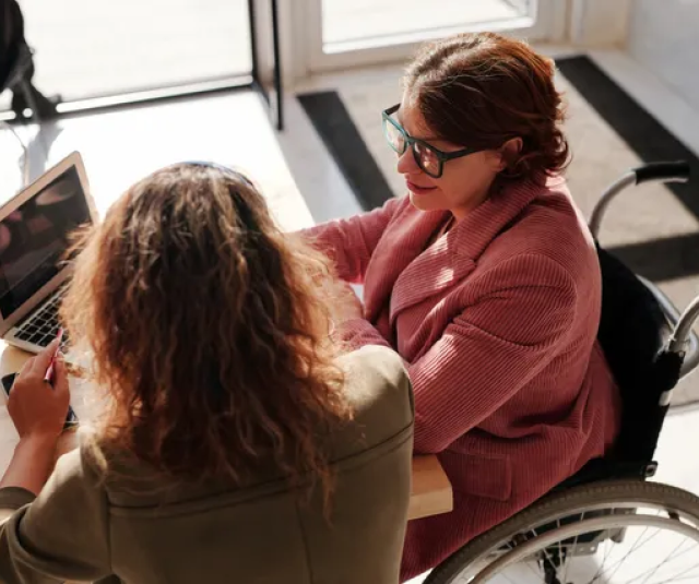 A woman in a wheelchair is sitting at a table using a laptop computer.