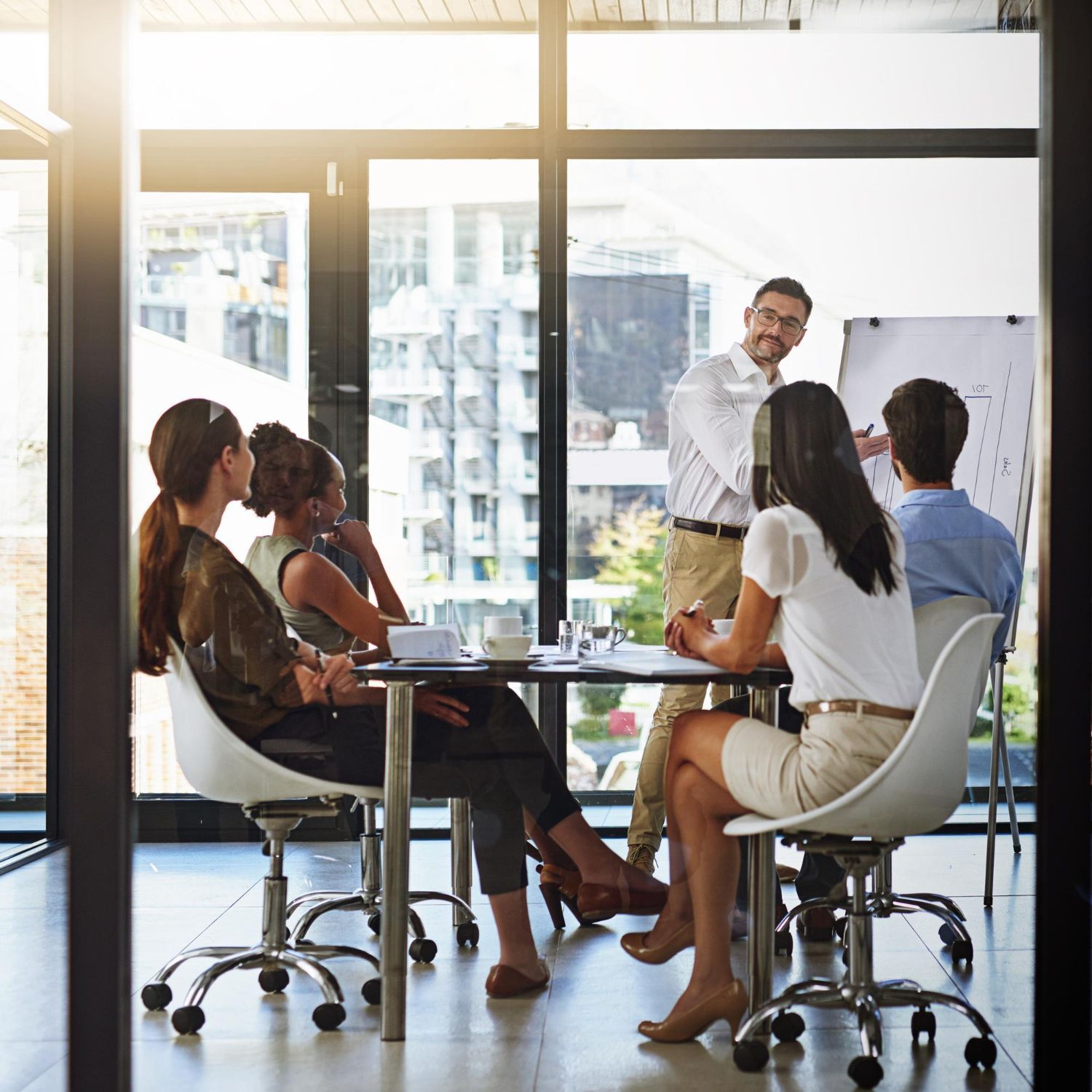 Office meeting: man presenting at whiteboard, five colleagues seated, city view.