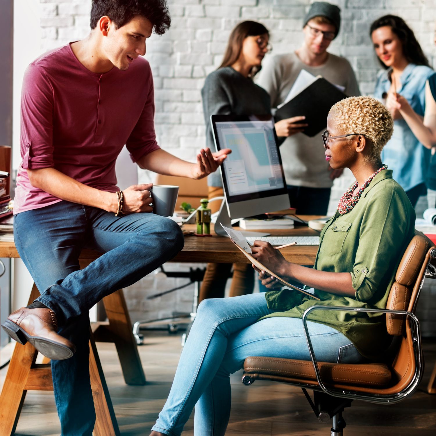 Group of diverse people in a modern office collaborating around a desk with a computer, discussing ideas and looking at documents.