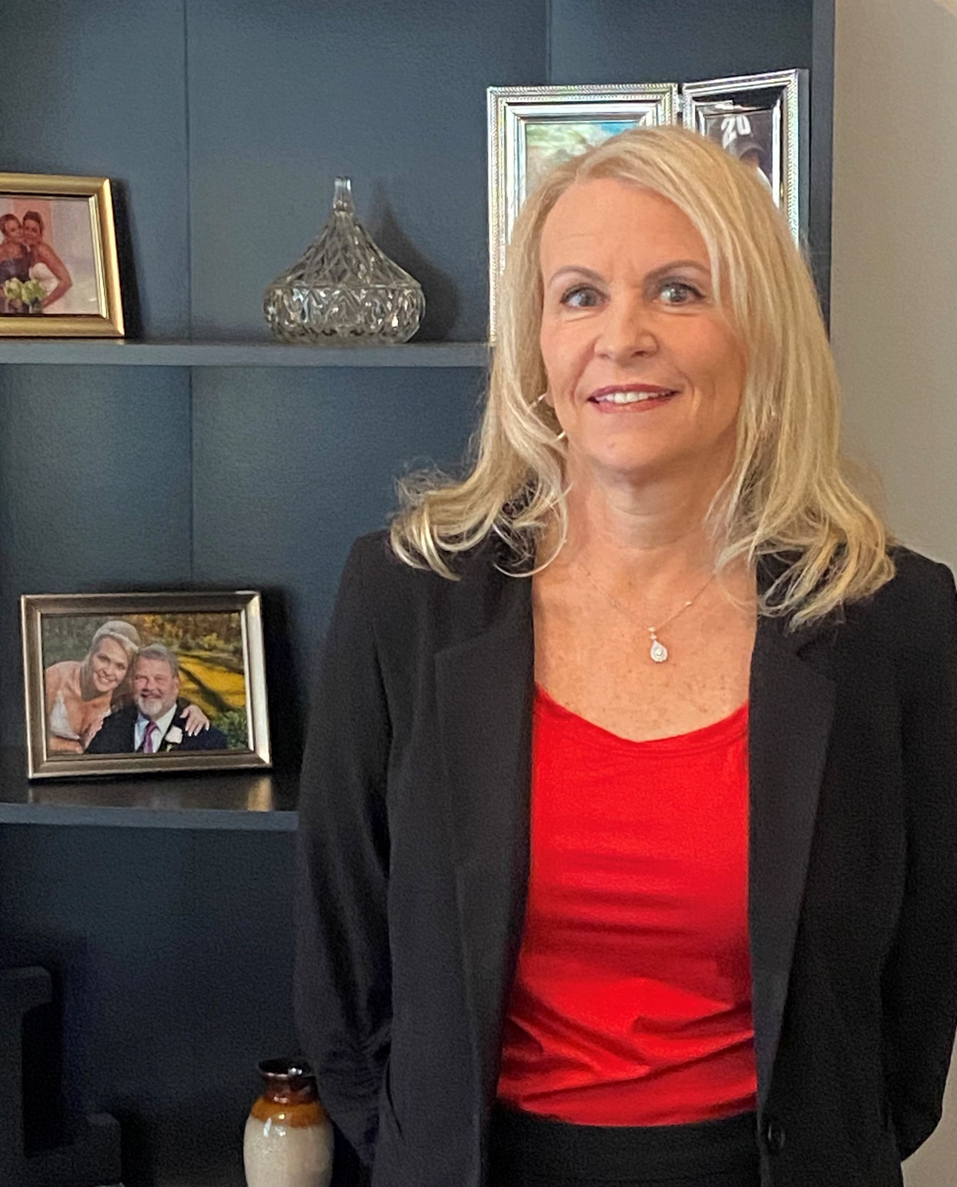 Blonde woman in red top and black blazer, standing by a shelf with photos, smiling.