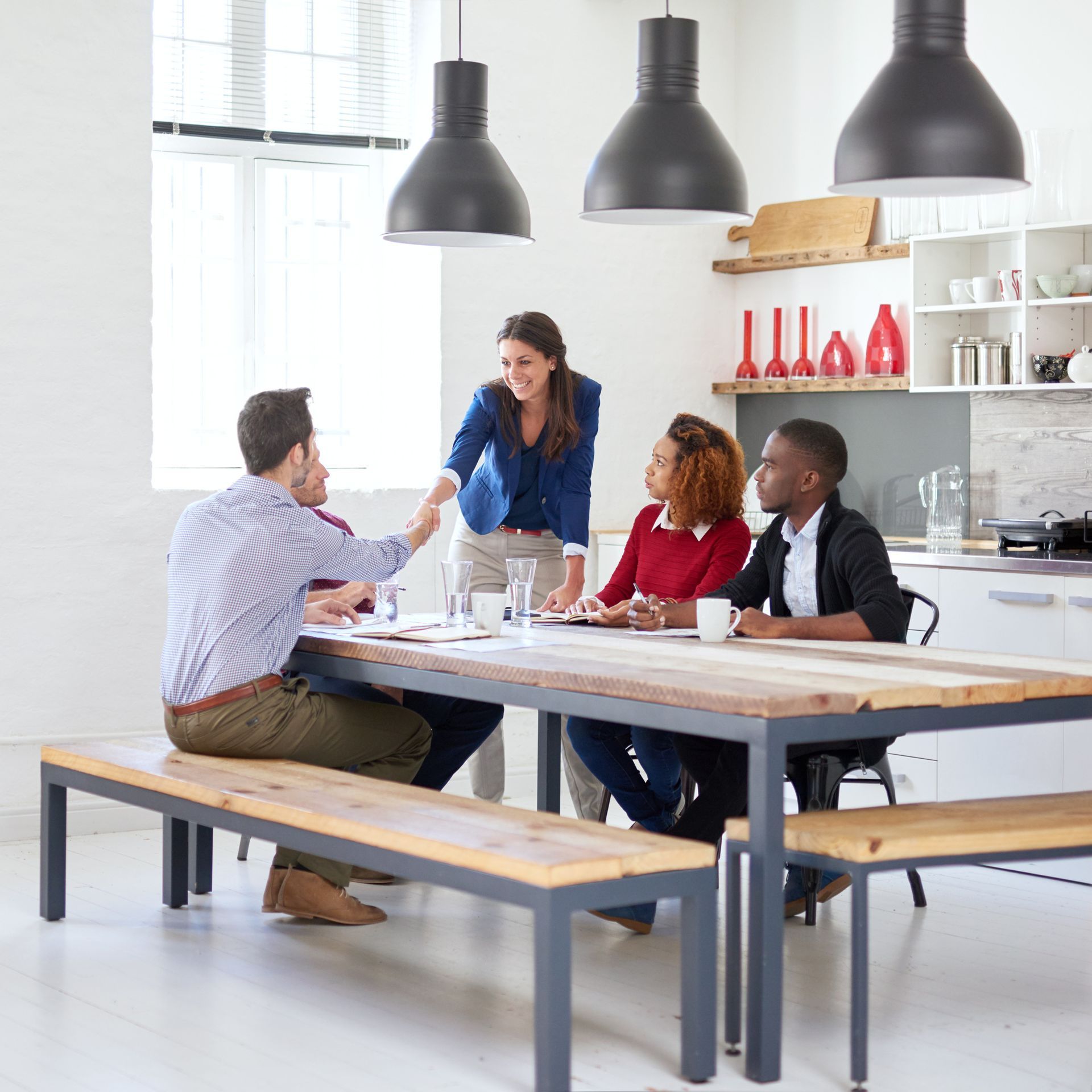 People at a table, one shaking hands. Bright interior with hanging lights, kitchen area visible.