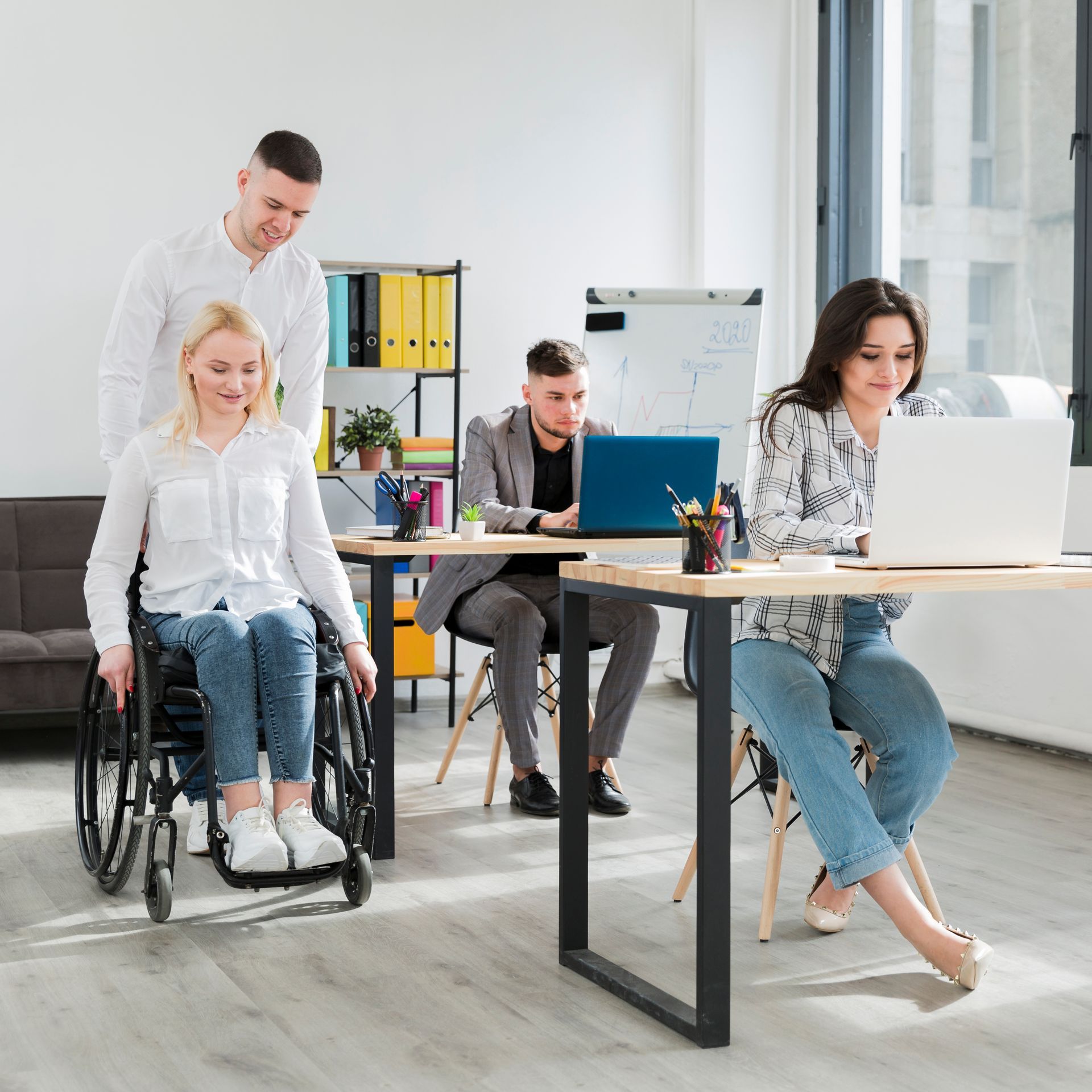 A woman in a wheelchair is sitting at a desk in an office with other people.